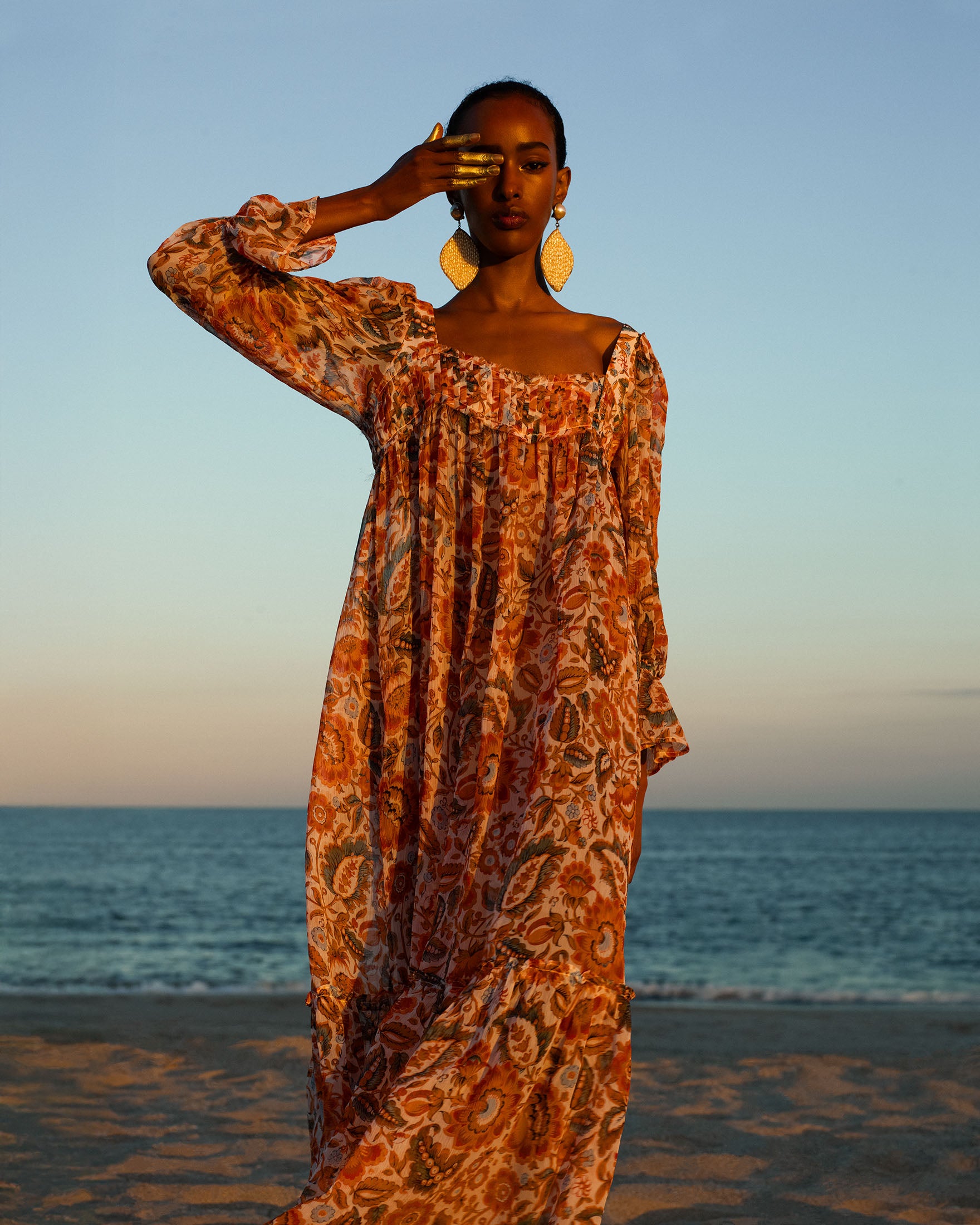 Wearing the Lilium Topaz Flower Dress, a person stands on the beach at sunset, raising one hand to shield their eyes.