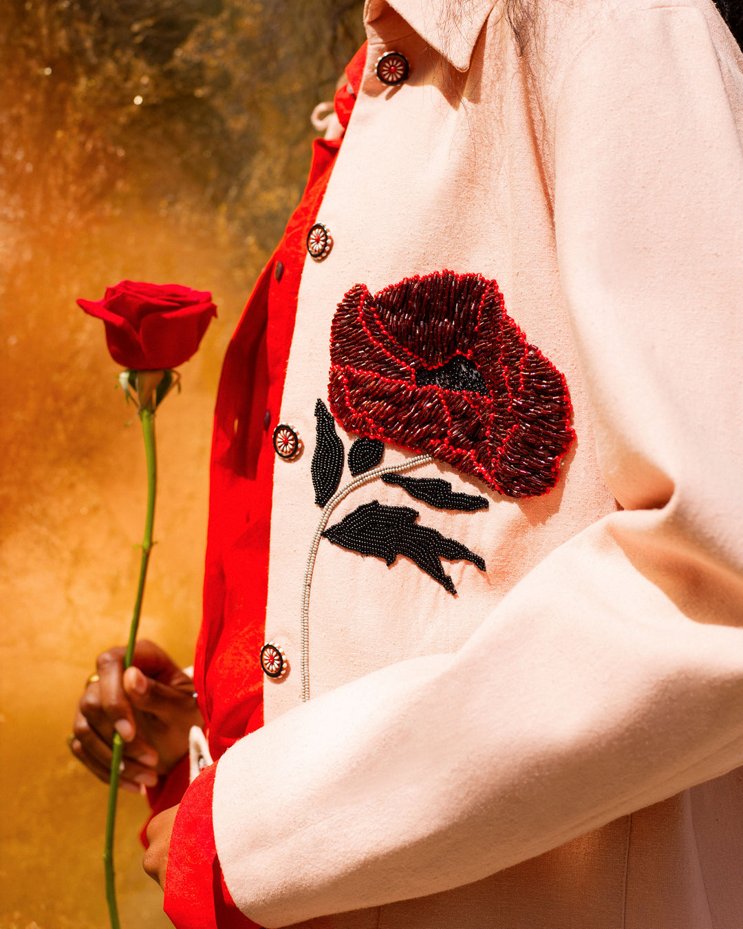 Wearing the Jude Ruby Poppy Jacket, adorned with a large red poppy, a person holds a single red rose against a warm golden background.