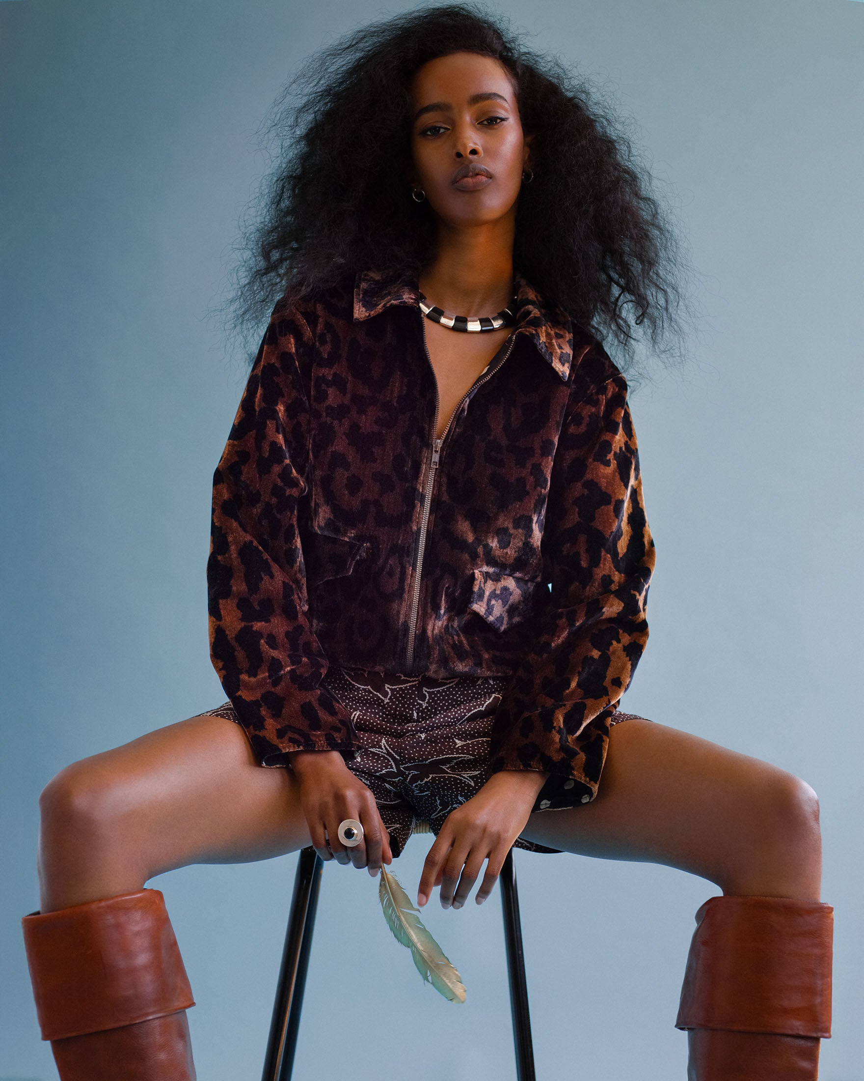Woman with voluminous hair sits on a stool against a blue background, wearing the Lex Leopard Velvet Jacket, printed shorts, brown knee-high boots, and holding a feather.