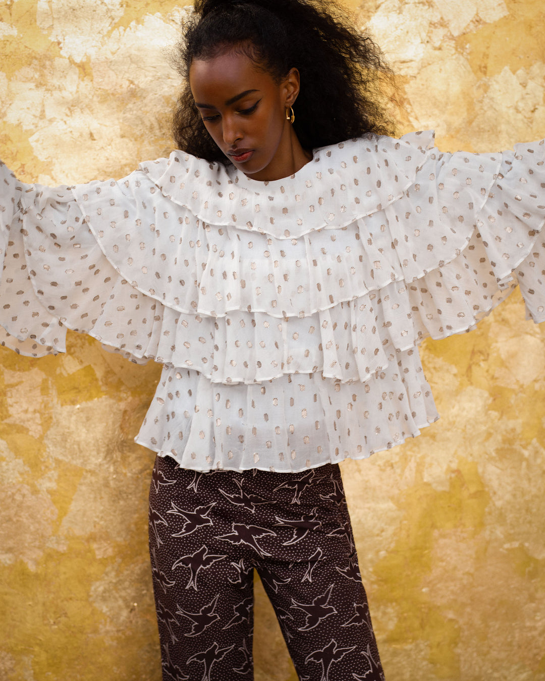 A woman stands against a gold textured wall wearing the Rufus Bronze Mist Top, a white blouse with ruffle details and wide sleeves, paired with dark bird-print pants.