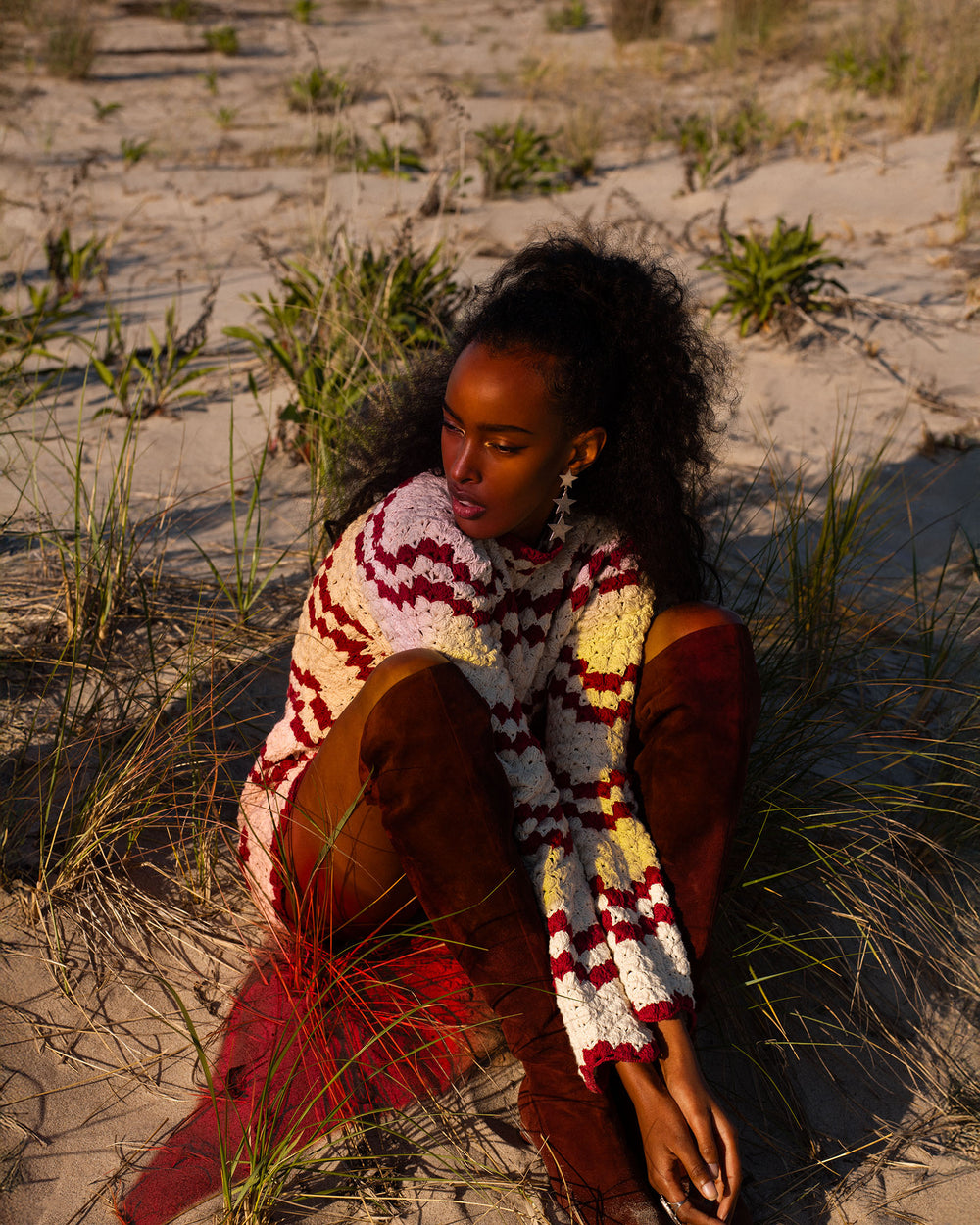 A person wearing a striped sweater and brown boots sits on sandy ground among grass, gazing dreamily to the side as if imagining the Romy Ruby Stripe Dress—the perfect holiday outfit with its delicate scalloped hem.