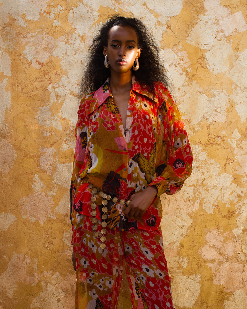 A woman stands against a textured yellow wall, wearing the Emery Marigold Meadow Shirt—vintage-inspired and airy in viscose-chiffon—paired with large earrings and loose, curly hair.