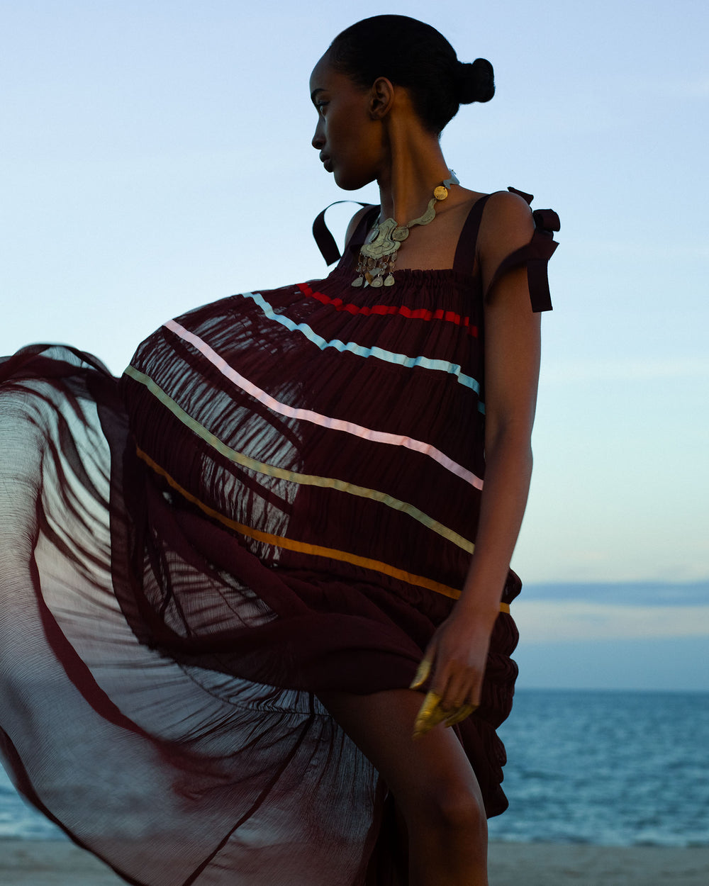 Standing on a sunset beach with ocean and sky behind, a person wears the Emrys Clove Aurora Dress—a flowing maroon maxi with horizontal colored stripes and ruched tiers—accessorized by a statement necklace.