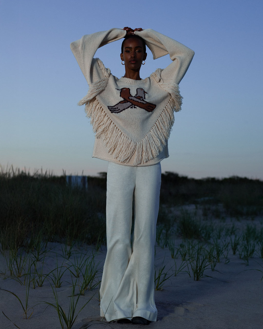 Standing on sandy grass at dusk, wearing cream wide-legged pants and the Rosa Lovebird Knit Sweater—a cozy baby alpaca crewneck with a bird design—arms raised overhead. The perfect winter layer.