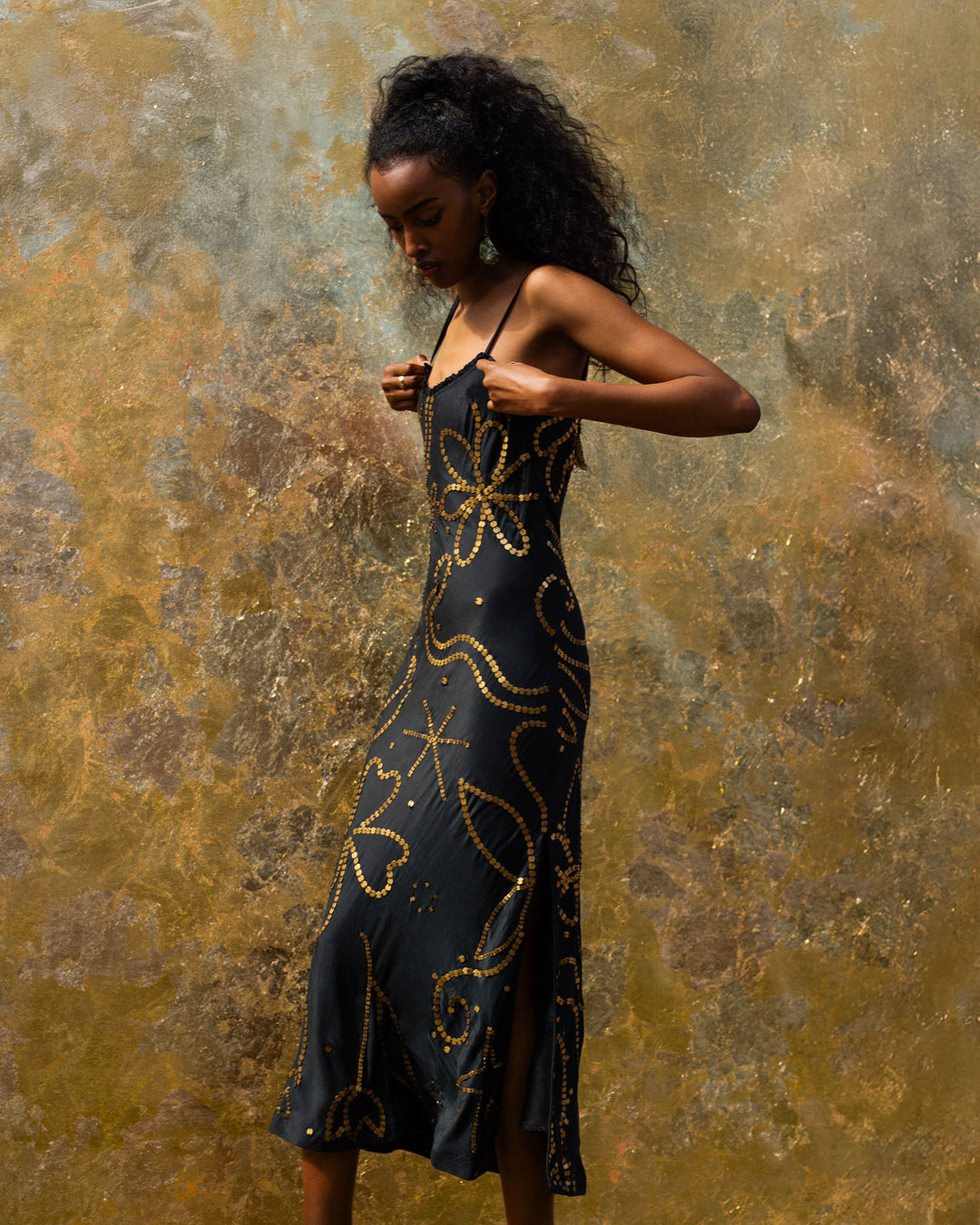 A woman in the Athens Jet Coin Silk Dress, featuring hand-loomed silk with gold patterns, stands against a textured gold background, holding one dress strap.