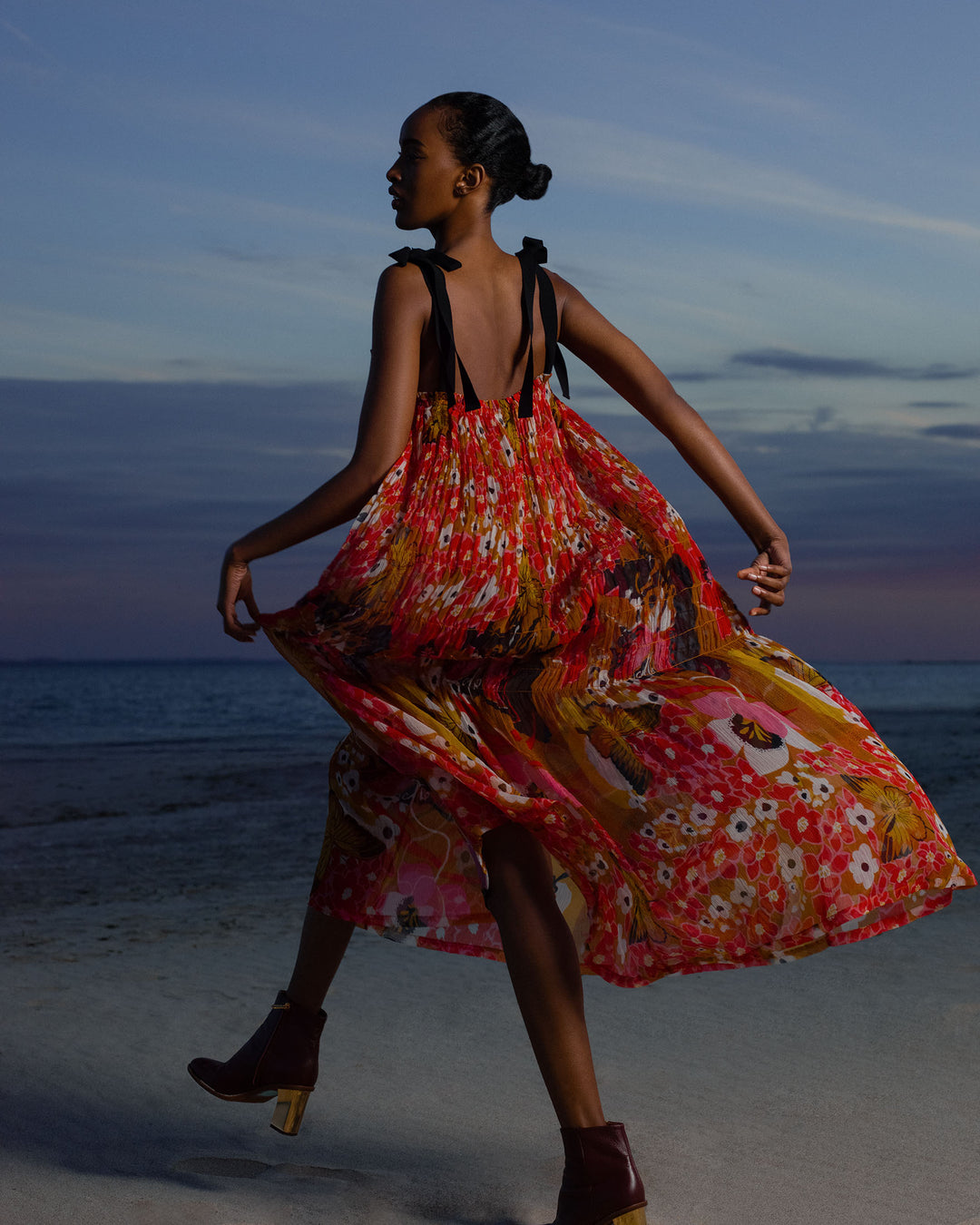 Wearing the Emrys Marigold Meadow Dress, a woman strolls along the beach at dusk, vibrant tiers of viscose-chiffon flowing as the ocean and a cloudy sky create a serene backdrop.