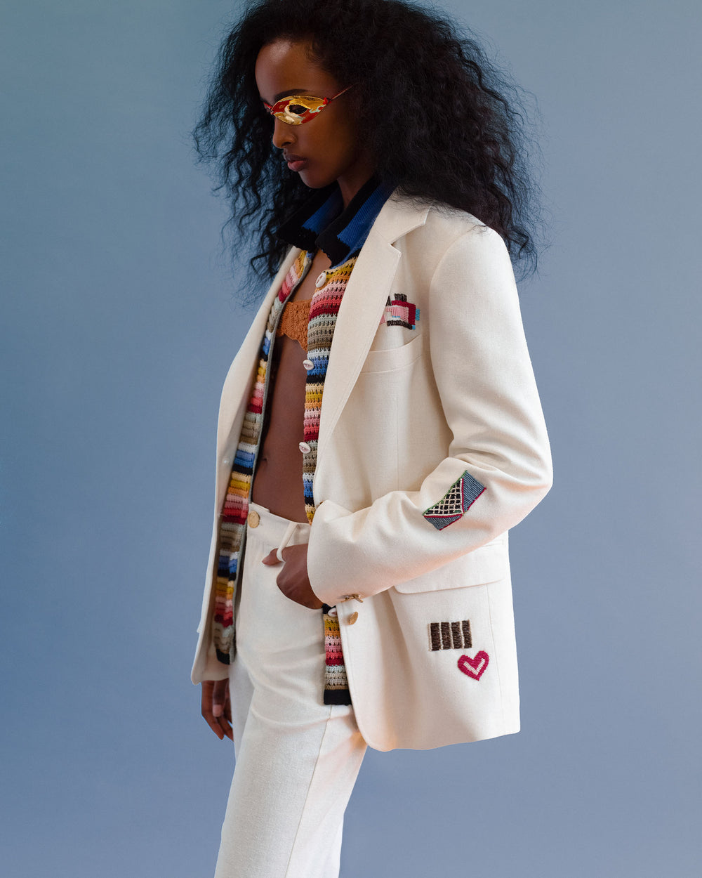 A woman wearing the Afternoon Abstract Tile Blazer, featuring white fabric with colorful embroidered patches, a multicolored crochet top, and yellow glasses stands against a blue background.