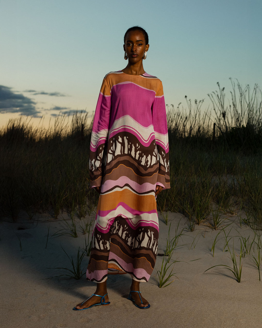 At dusk, a person stands on sandy ground framed by tall grass and a cloudy sky, wearing the Niall Bloomshade Dress—a maxi dress with flared sleeves and a pink, orange, brown, and white abstract print.
