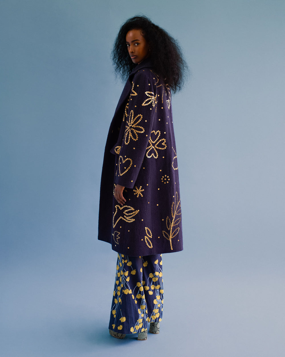 A woman with long, curly hair stands sideways against a light blue background, wearing the Petunia Midnight Coin Coat—a navy Khadi cotton coat with yellow patterns—paired with blue pants decorated in yellow designs.