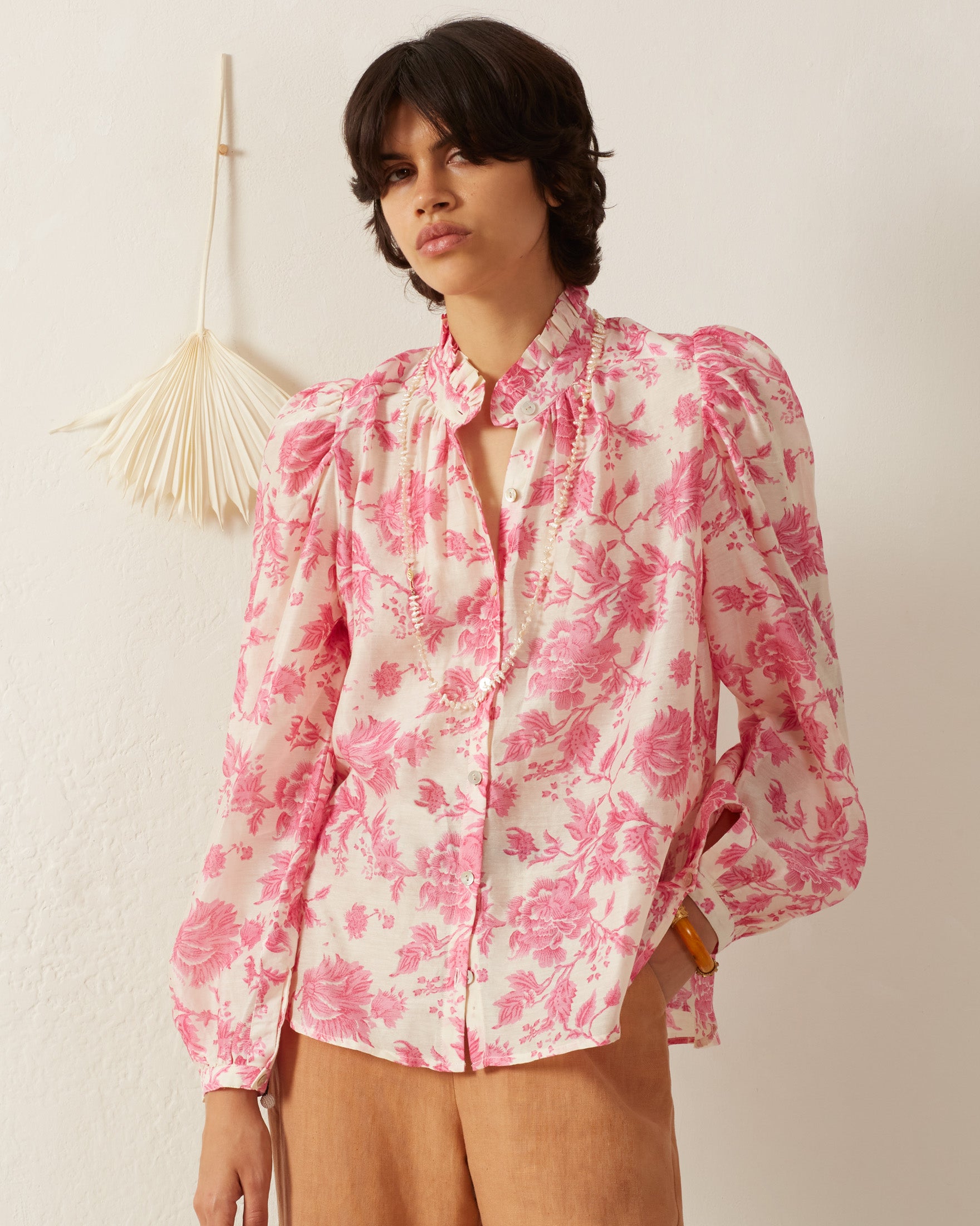 A person with short, dark hair is wearing a pink floral blouse with long sleeves and beige pants, standing against a light-colored wall with a decorative hanging.