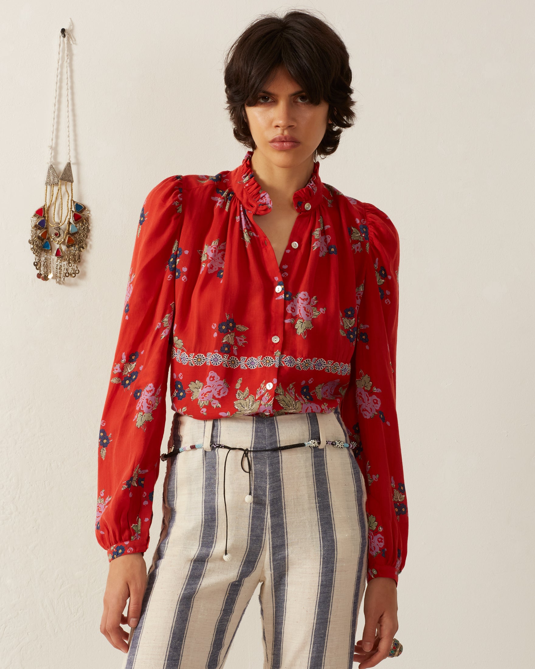 A person with short, dark hair is wearing an Annabel Carmine Bouquet Shirt, which features a vintage-inspired red floral print, paired with high-waisted striped pants. They stand against a plain wall with a decorative necklace hanging in the background.