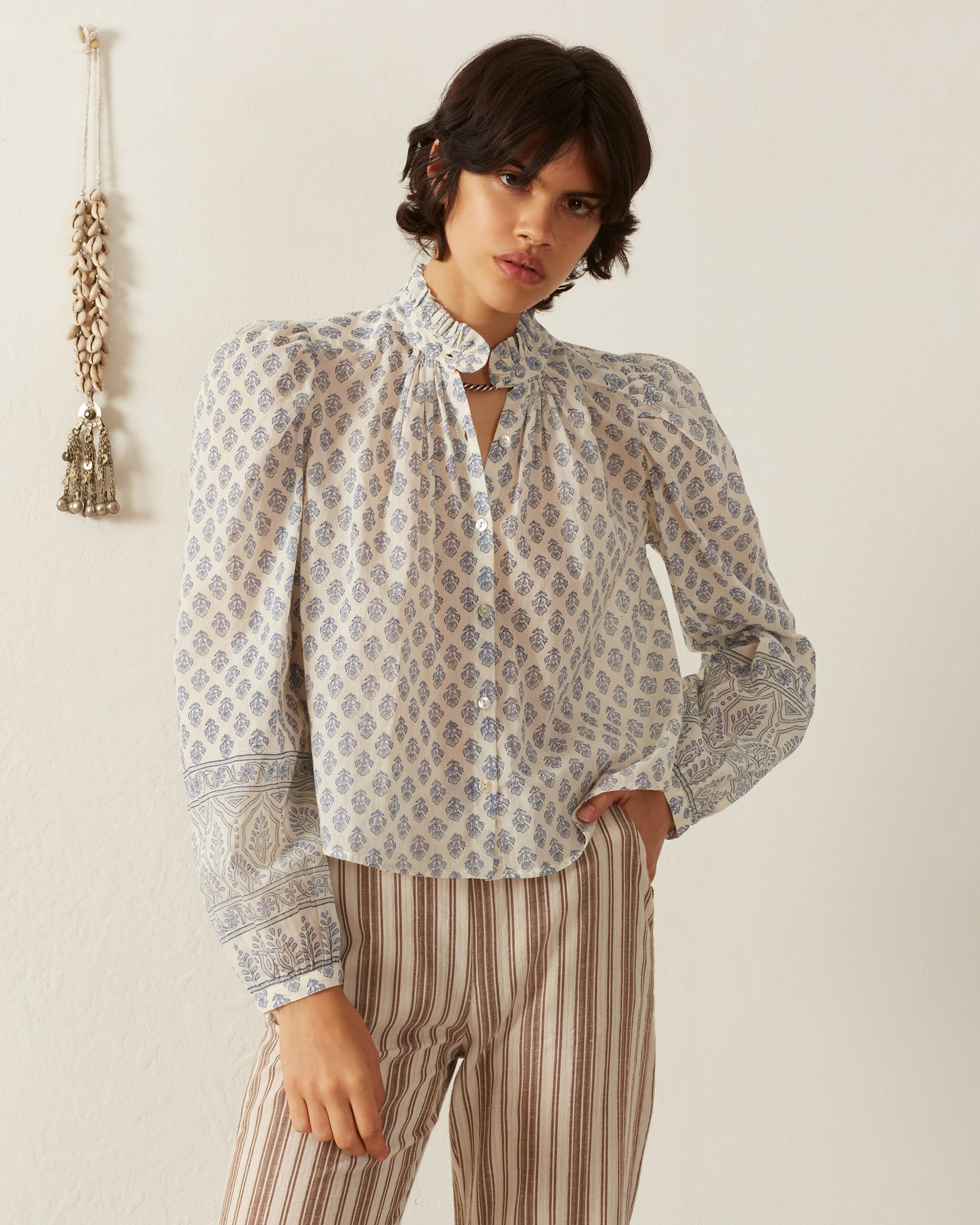 A person with short dark hair wearing the vintage-inspired Annabel Delft Bhutti Shirt, which features a blue floral print, and beige striped pants stands against a cream background. A decorative tassel is seen hanging on the wall.