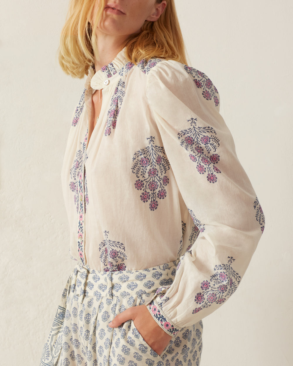 Wearing the Annabel Freesia Phlox Shirt, a person stands against a plain background. The light, vintage-inspired cotton voile blouse is adorned with pink and purple floral designs, paired with matching pants that showcase a blue floral pattern. Their blonde hair is down, partially obscuring their face.