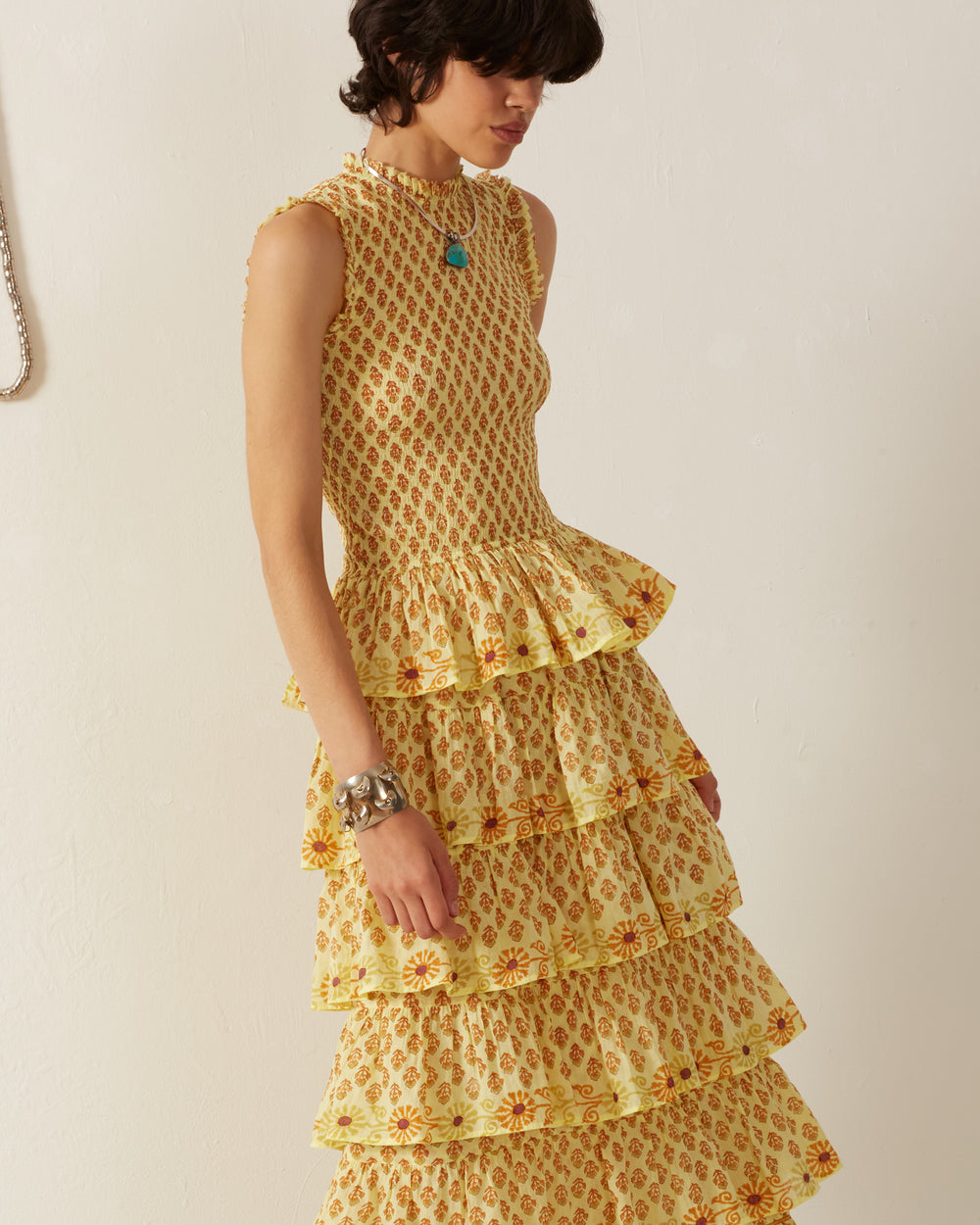 A person with short, dark hair wears the Cabana Saffron Bhutti Dress, a sleeveless, ankle-length cotton voile dress adorned with a vibrant red floral pattern and multiple ruffle tiers. They accessorize with a turquoise necklace and a large silver bracelet.