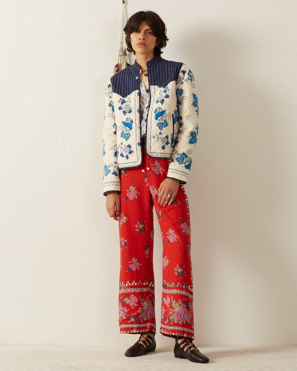 A person stands against a white wall wearing the Claude Blue China Panel Jacket, which features a quilted floral print and a band collar, paired with red patterned pants. They have mid-length dark hair and are wearing black shoes.