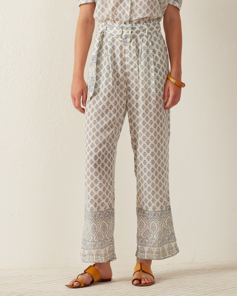 A person is seen wearing the Colette Delft Bhutti Pant, featuring a blue floral print and a wide-leg design, while standing against a neutral background. The high-waisted pants, made from Tencel fabric, are fashionably paired with open-toe sandals and accessorized with a bracelet on their left wrist.