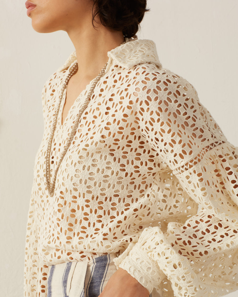 A person wearing the Dylan Clover Eyelet Top, a vintage-inspired cream-colored cotton blouse with a floral pattern, stands against a light background. The top features a collar and long sleeves and is elegantly accessorized with a long, delicate necklace, exuding timeless charm.