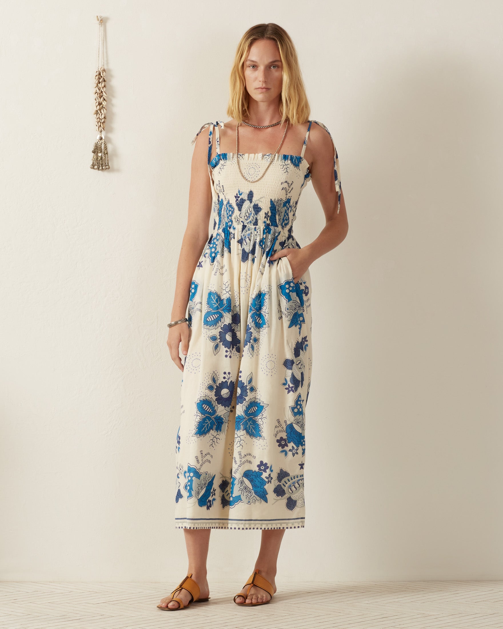 A woman in the Kaia Blue China Panel Dress, with its tie straps and smocked bodice, reminiscent of vintage Provençal textiles, stands against a plain wall. She wears brown sandals and bracelets, her hands nestled in her dress pockets as a decorative tassel adorns the wall.