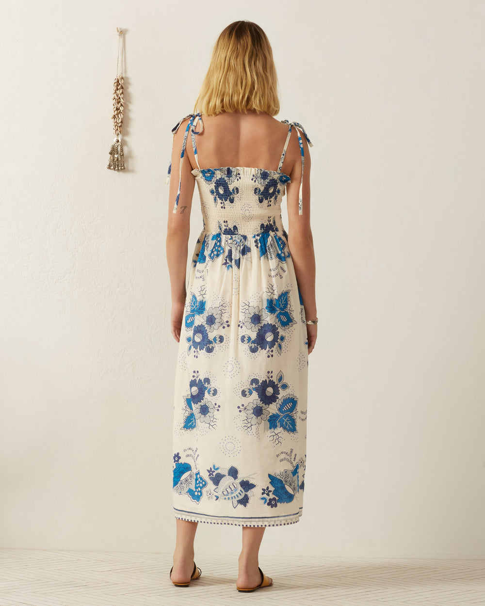 A woman stands with her back to the camera, wearing the Kaia Blue China Panel Dress, a long, sleeveless piece inspired by vintage Provençal textiles. Her blond hair is loose, and she is barefoot on a light wood floor. A decorative hanging adorns the off-white wall.