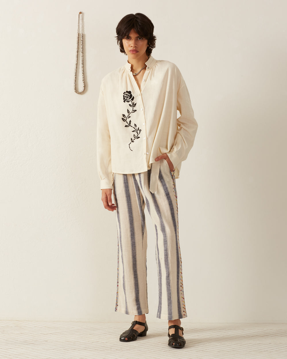 A person with medium-length hair stands against a plain wall, wearing the cream-colored Kiki Noir Rose Embroidered Shirt paired with striped pants. A necklace hangs on the wall beside them, and they are wearing dark sandals made of fine Khadi cotton.