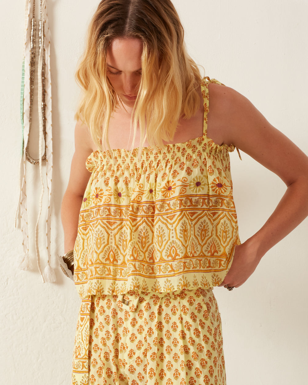 A woman wearing the Lila Saffron Bhutti Top, a block-printed yellow and orange sleeveless top paired with matching pants, stands against a white wall. Her face is framed by her blonde, wavy hair as she looks down. A beaded necklace enhances the background wall, adding subtle charm to the cotton voile ensemble.