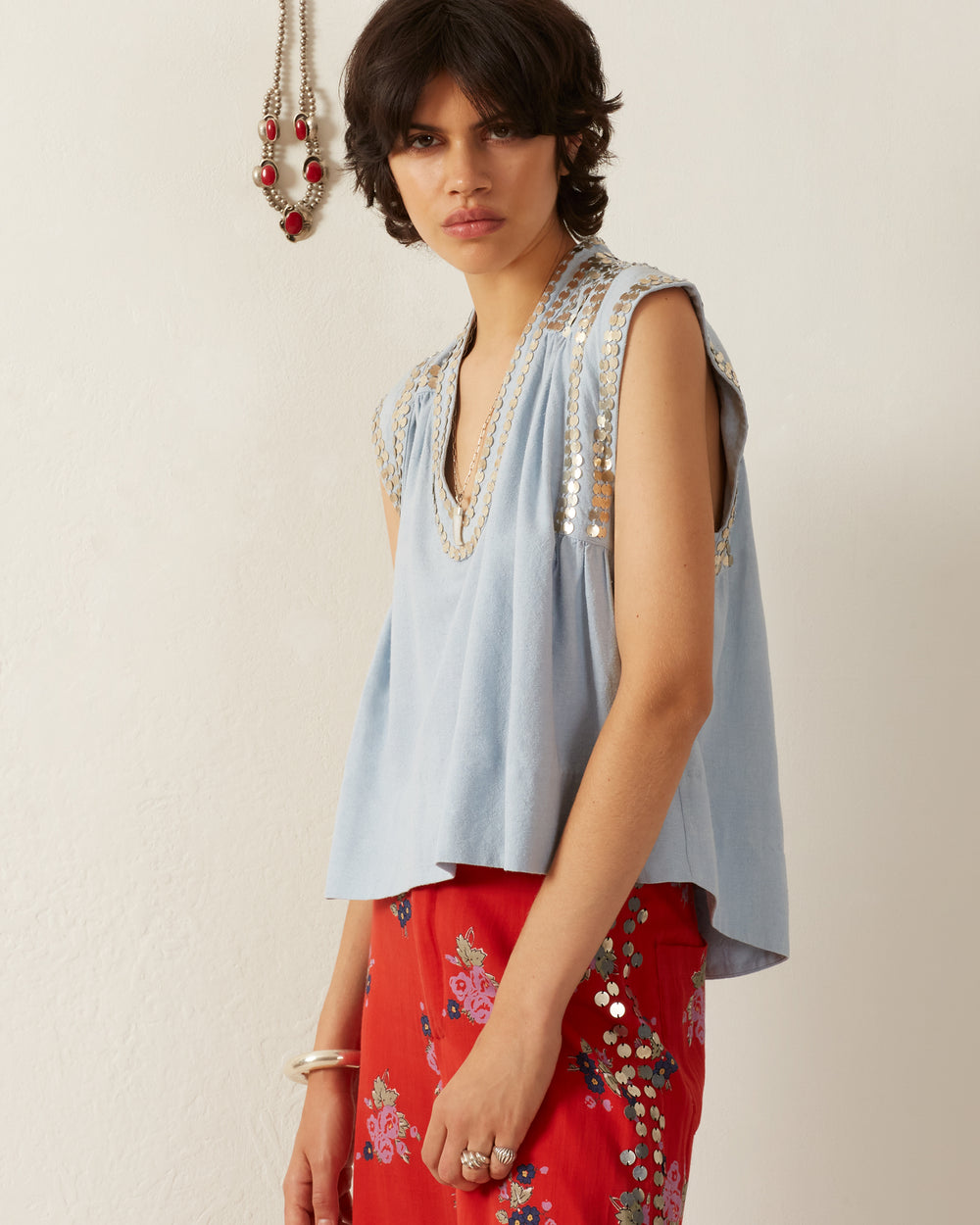 A person with short dark hair is seen wearing the Nehru Silver Sky Top, a light blue garment featuring hand-embellished decorative details, paired with a red floral-patterned skirt. In the background of the minimal, light-colored room hangs a beaded necklace on the wall.