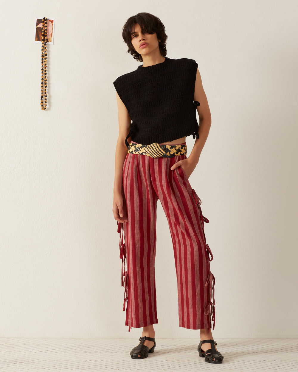 A person with shoulder-length dark hair stands against an off-white wall, embodying a '70s vintage-inspired style. They wear the Oona Onyx Crochet Top, paired with red and beige striped pants tied at the waist and sandals. A hand-crocheted patterned belt adds charm, while a small wall decoration is visible above.