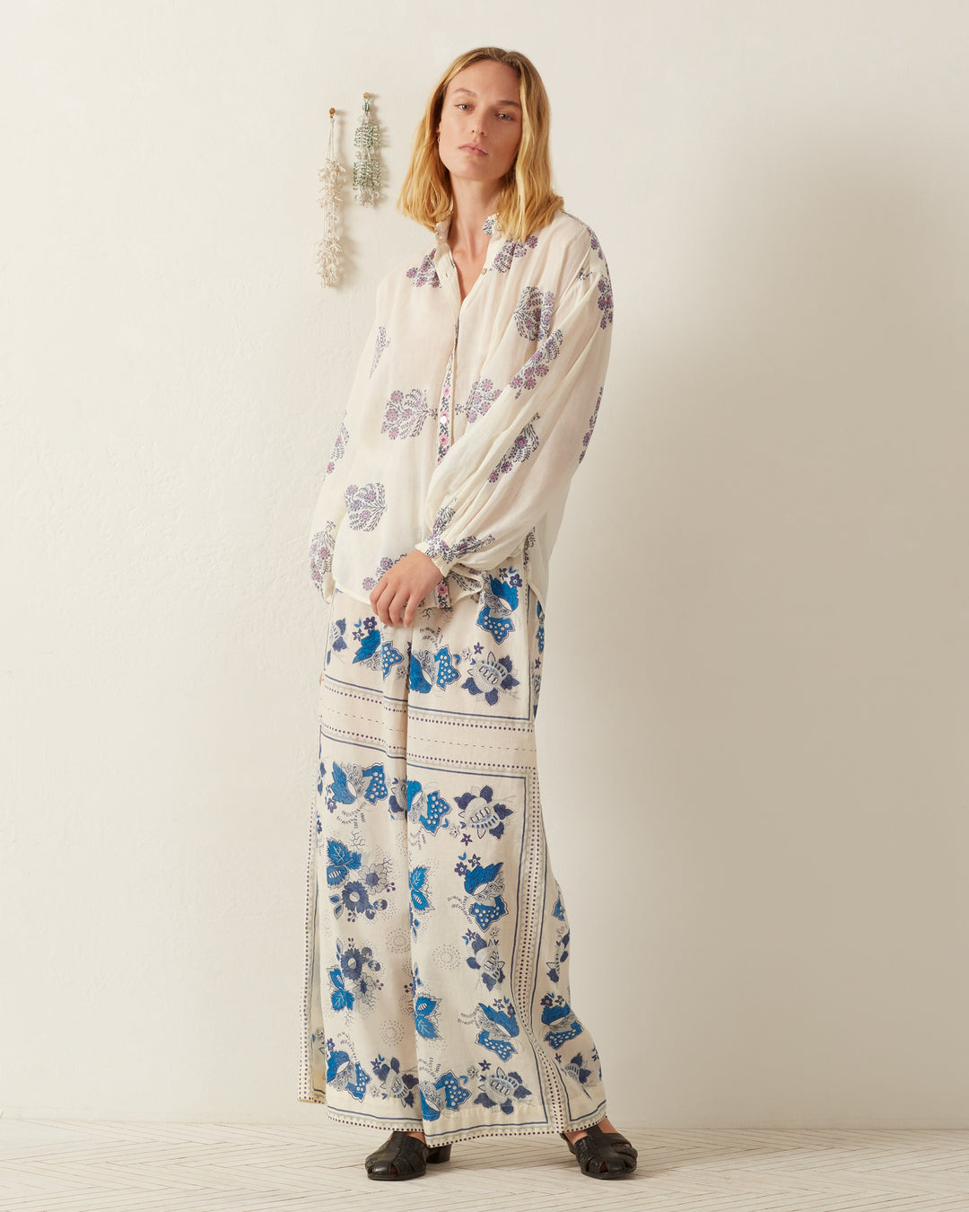 In a white room, a person stands wearing the flowy Poet Freesia Phlox Blouse paired with wide-leg pants, both adorned with blue floral designs. Their shoulder-length hair complements the black shoes they wear as dried flowers hang artfully on the wall in the background.