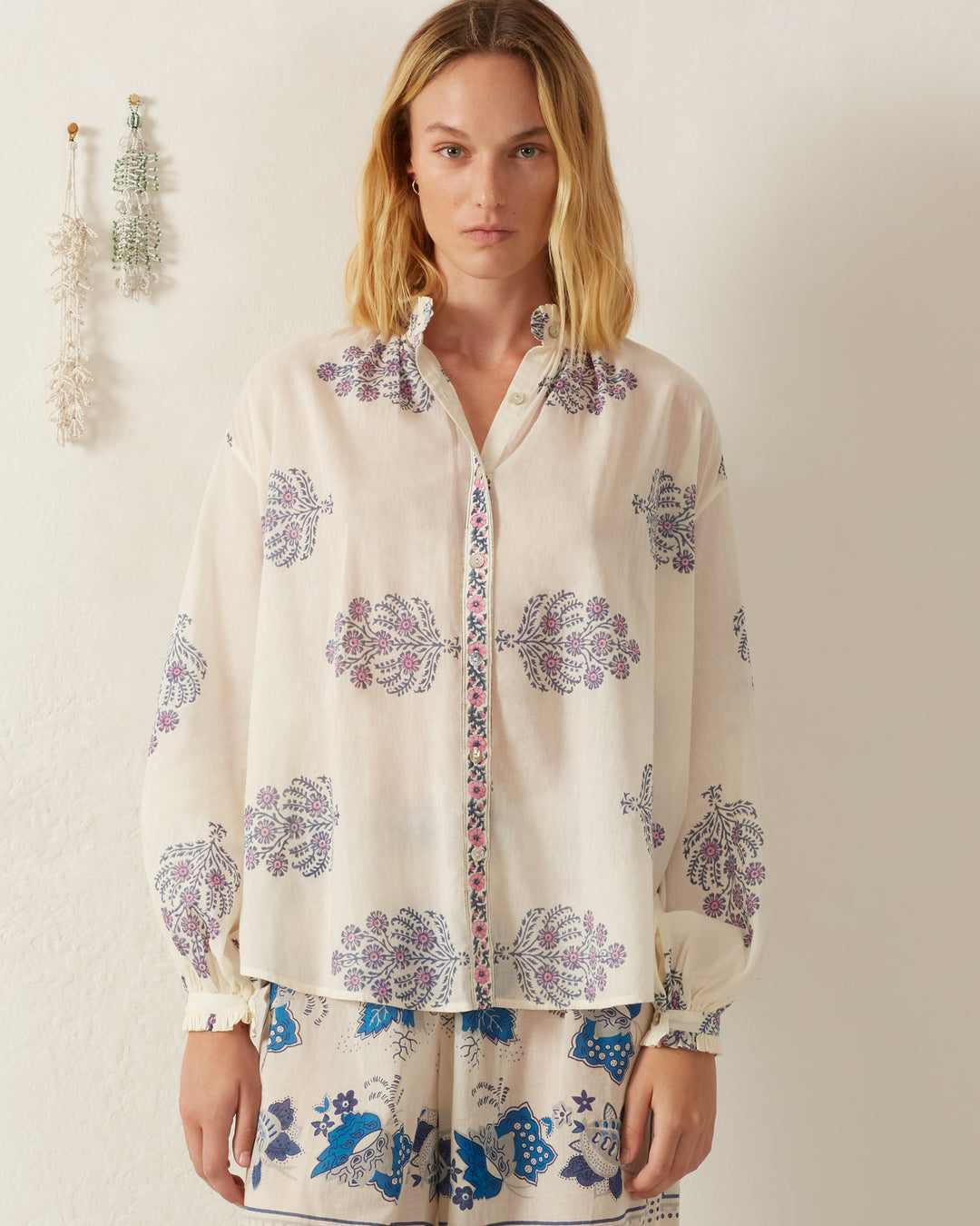 A woman with a neutral expression stands against a light background wearing the Poet Freesia Phlox Blouse, which features purple floral patterns that give it a vintage-inspired folk shirt look, paired with matching shorts. Her hair is down, and decorative hanging pieces adorn the wall beside her.