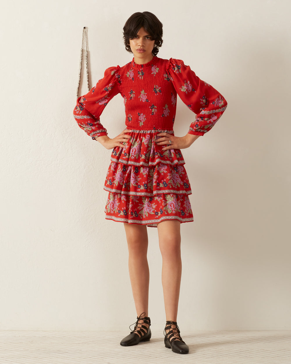 Indoors, a person showcases the Selma Carmine Bouquet Dress, which features a red floral print with puffed sleeves and a tiered skirt that evokes a vintage-inspired silhouette. They pair it with black lace-up shoes and pose against a plain white wall, while a small chain accessory hangs in the background.