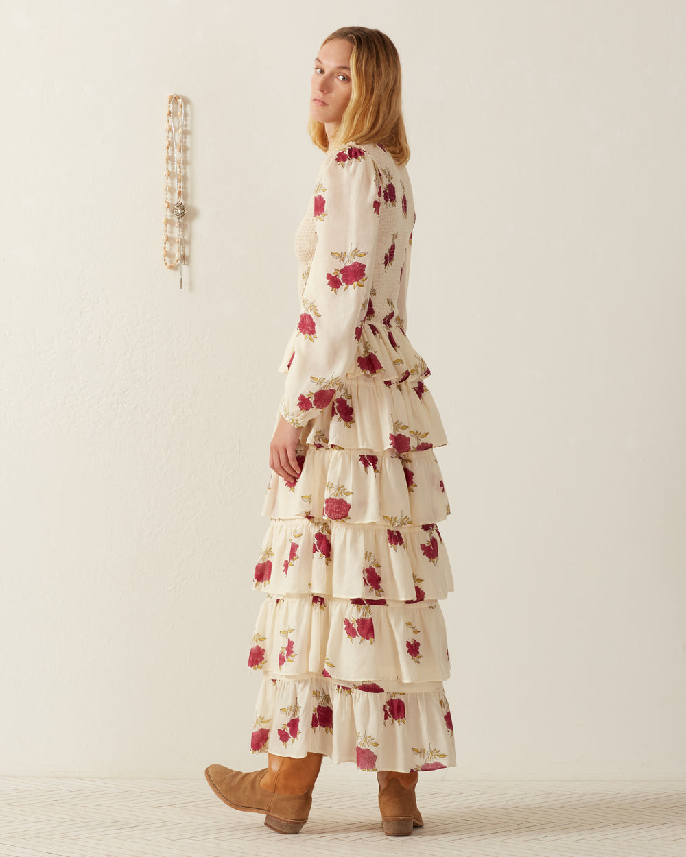A person with long hair stands sideways, dressed in the Shelby Cerise Rosette Dress paired with brown boots. The background features a light-colored wall with bead decoration.