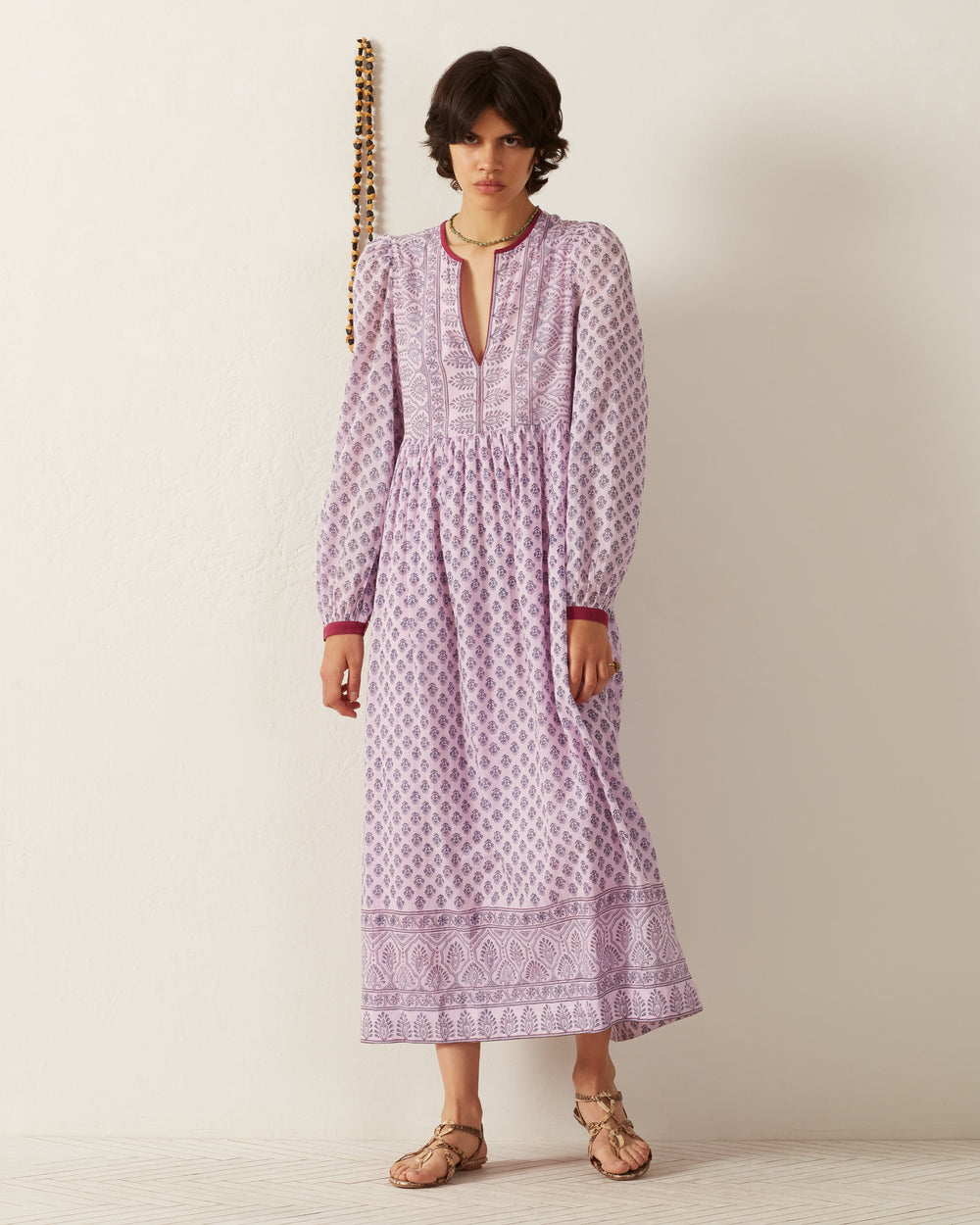 A person in a Winifred Lilac Bhutti dress, featuring a lively floral print and V-neckline, stands in front of a plain white wall. They wear sandals and maintain a neutral expression. Beside them, a beaded decor adds an artistic touch to the scene.