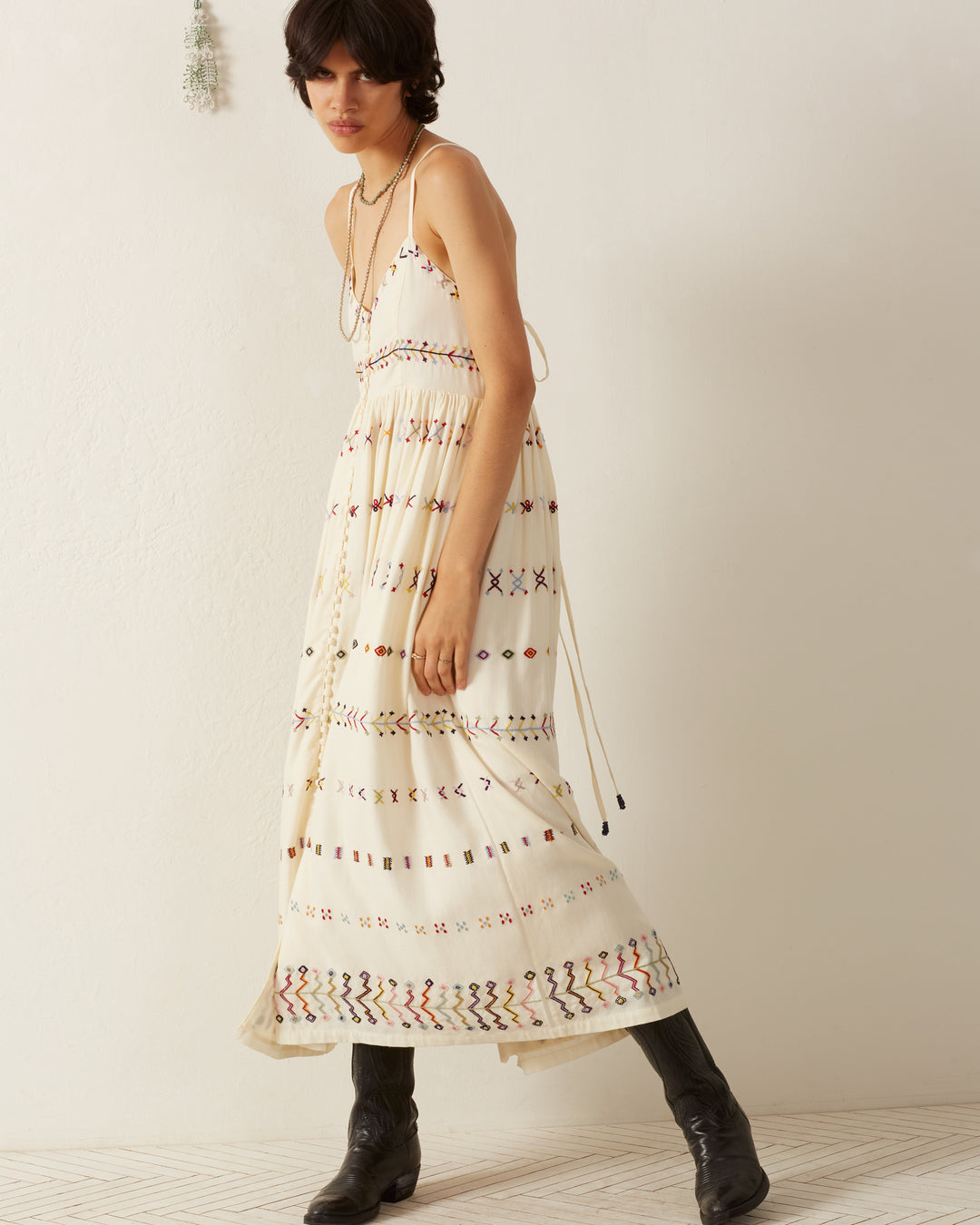 A person wearing the Skylar Aurora Arrow Dress, a long ivory sleeveless piece featuring colorful geometric patterns, stands against a plain white wall. The hand-embroidered details add charm to their short dark hair and black boots.