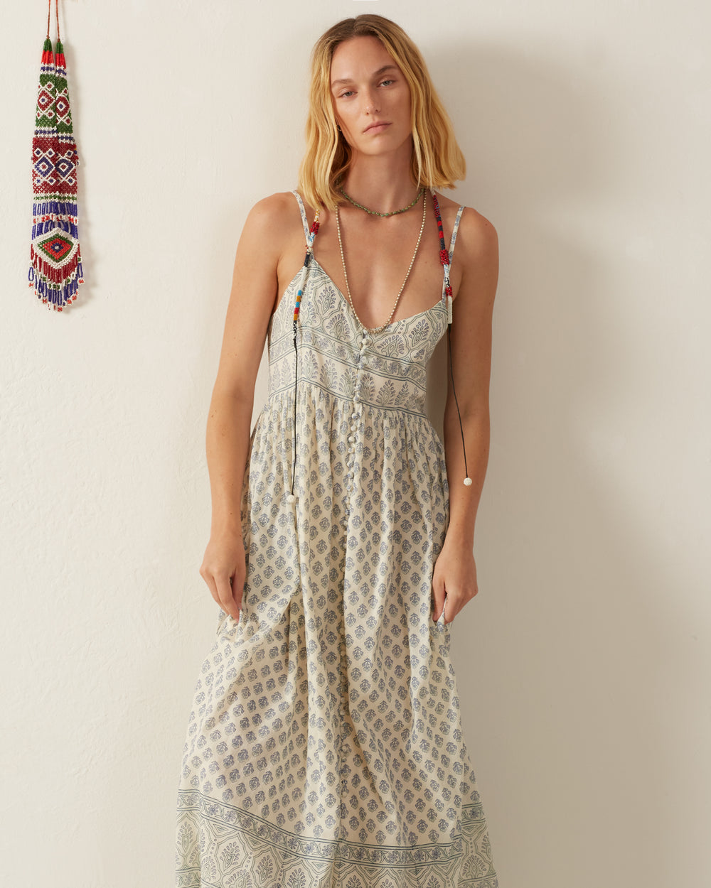 A person wearing a Skylar Delft Bhutti Dress stands against a plain light-colored wall. They have long hair and are accessorized with a beaded necklace. A colorful, hanging beaded decoration is visible on the left side of the image.