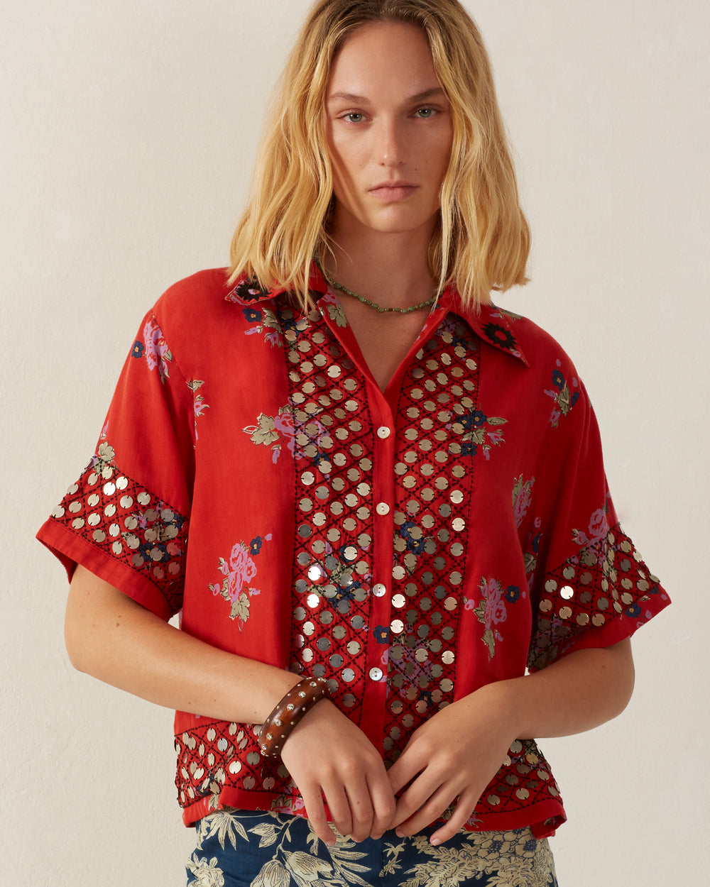 A person with shoulder-length blonde hair is wearing the Stevie Carmine Coin Shirt featuring vintage Hawaiian floral patterns and mesh detailing. With a neutral expression, they stand against a plain backdrop, while hand-embroidered beadwork on their necklace and bracelet completes the look.