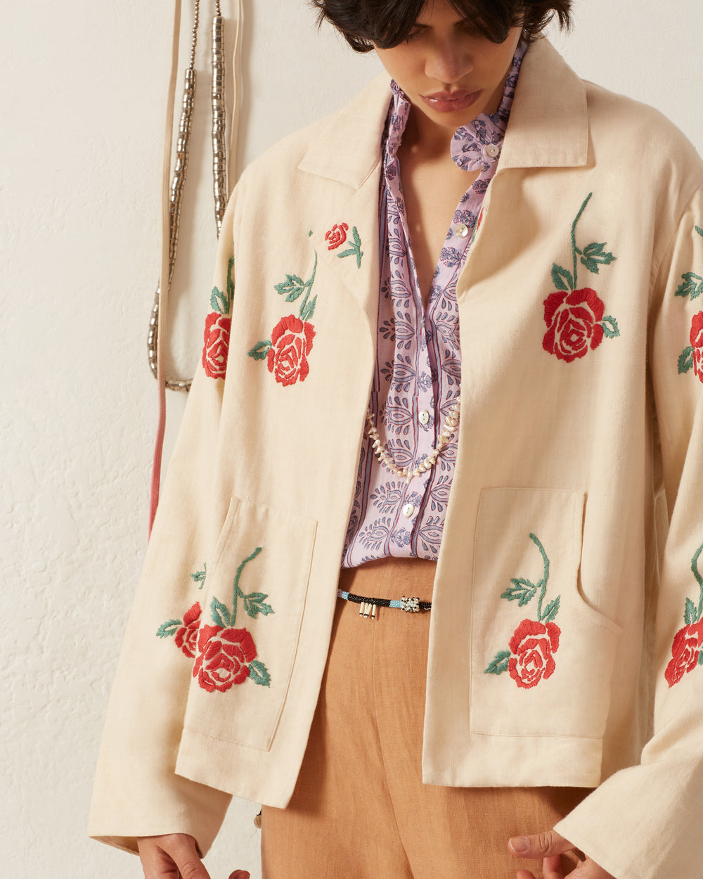 A person wearing the Western Blush Rose Jacket with hand-embroidered red roses over a patterned purple shirt pairs the outfit with brown pants and a belt, adorned with visible necklaces. The background features a white wall and hanging accessories.