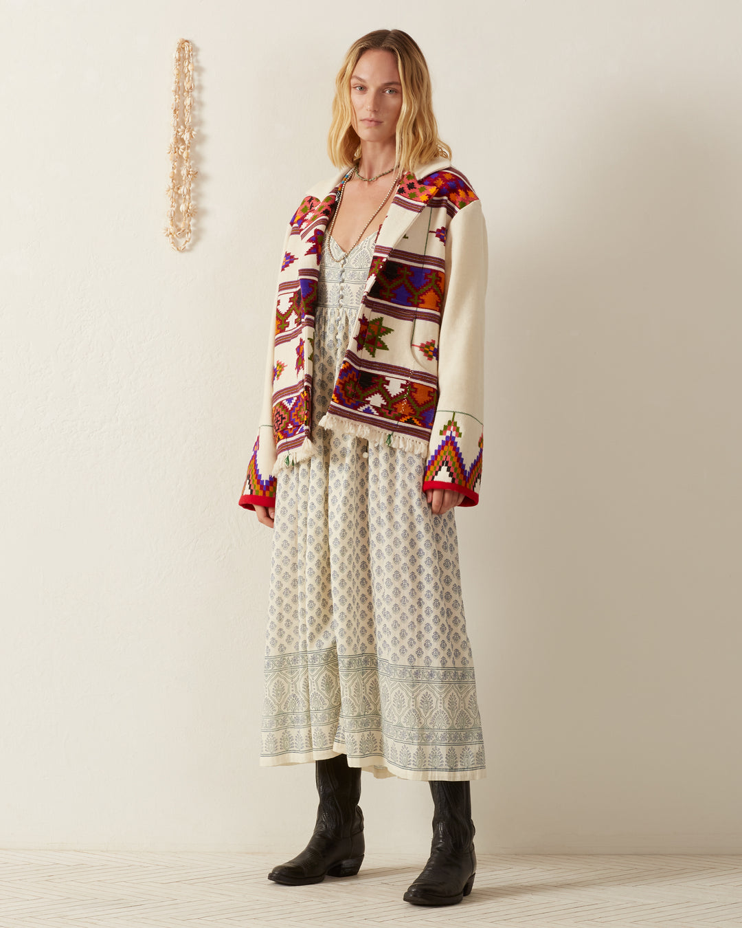 A person stands against a light-colored wall, wearing a long patterned dress and the Western Tiger Eye Blanket Jacket with black boots. They look forward with a relaxed expression, while a string of decorative beads hangs on the wall beside them.