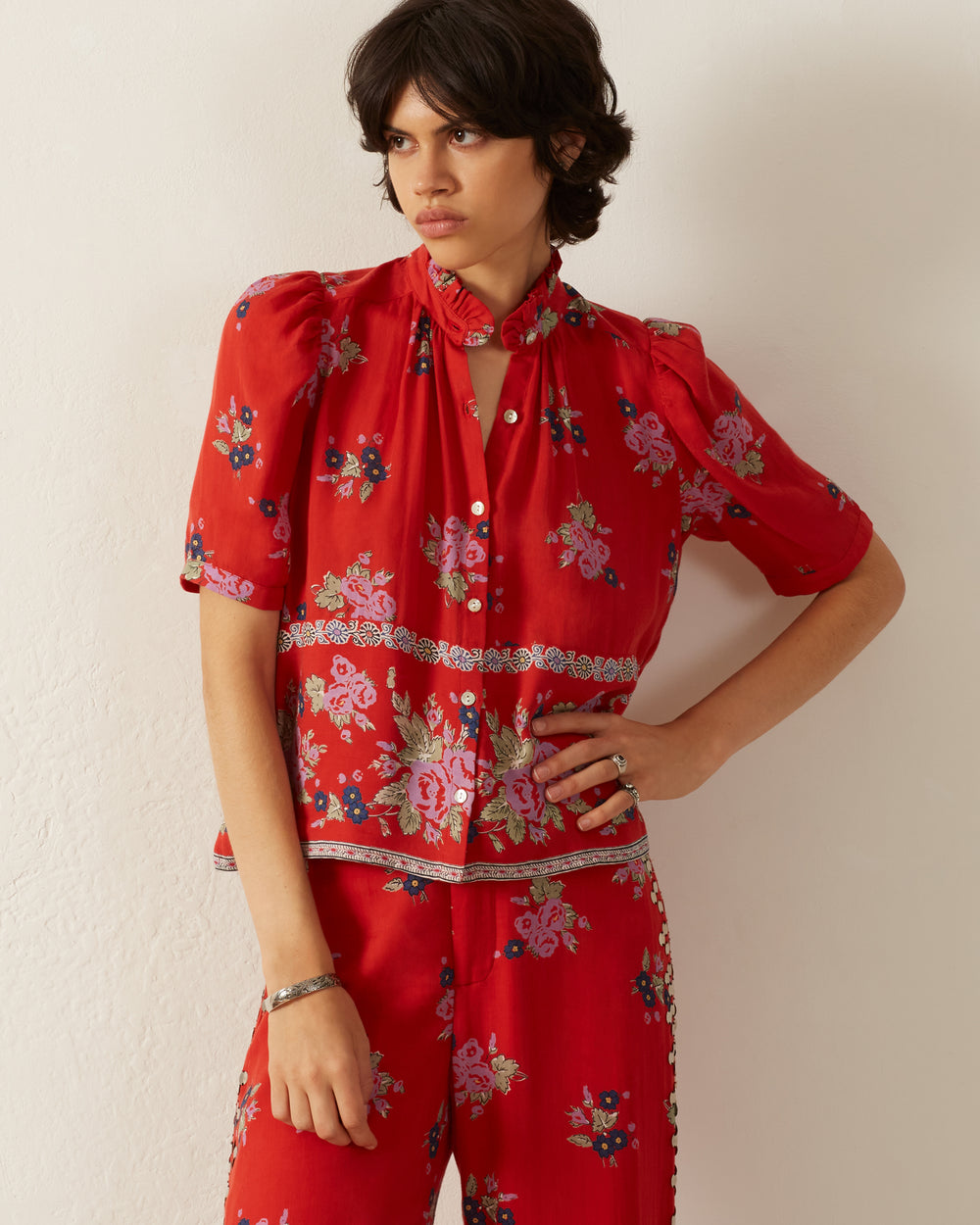 A person with short dark hair stands against a white wall wearing the vibrant red Winnie Carmine Bouquet Shirt with its vintage-inspired floral print. The shirt features short sleeves, a high collar, and button details. They have a neutral expression with one hand resting on their hip.