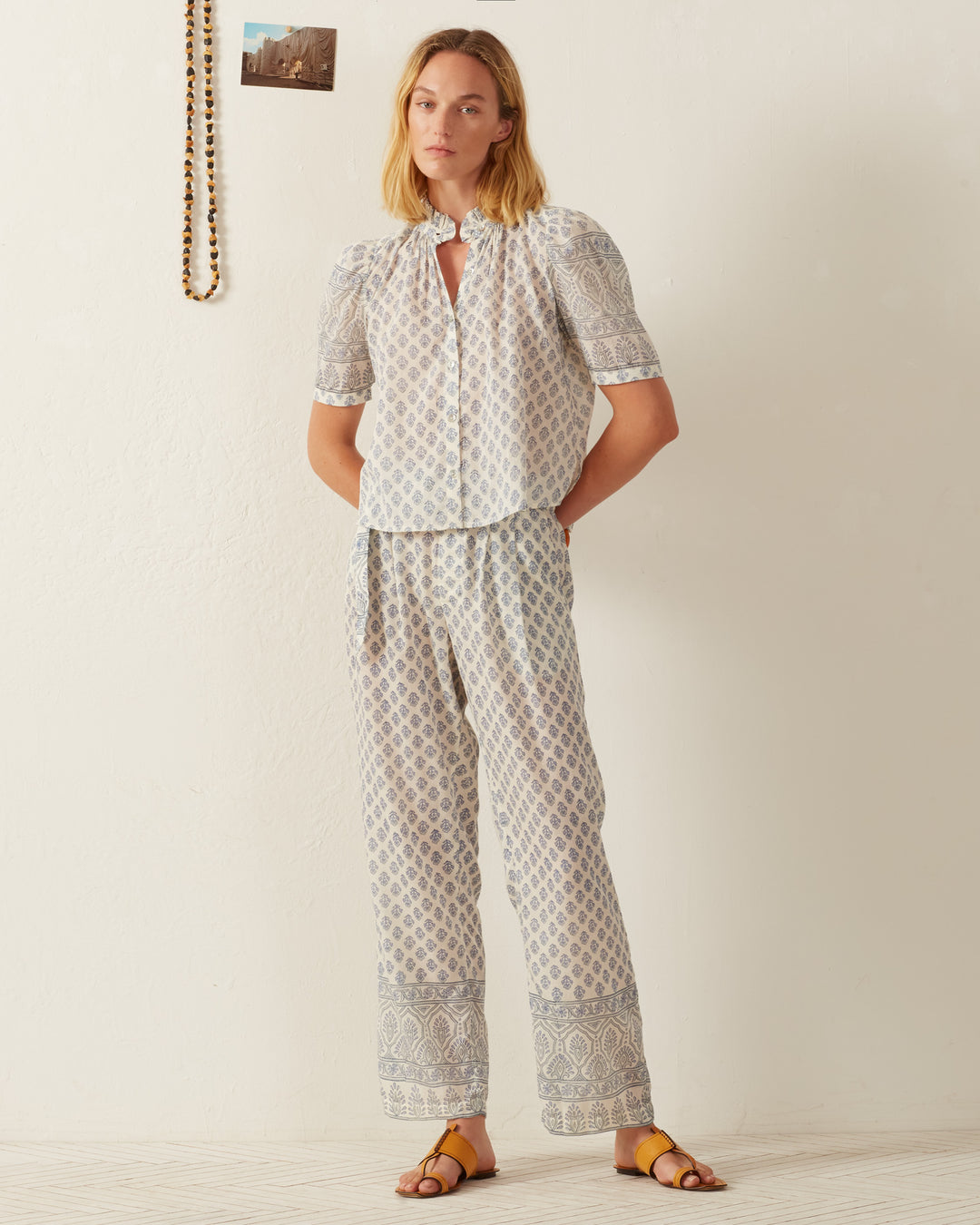 A woman stands against a white wall wearing the Winnie Delft Bhutti Shirt, a vintage-inspired piece with a blue floral print, paired with matching pants. She is barefoot on a light wooden floor. A string of beads and a small photo are hung on the wall beside her, adding charm to the serene setting.