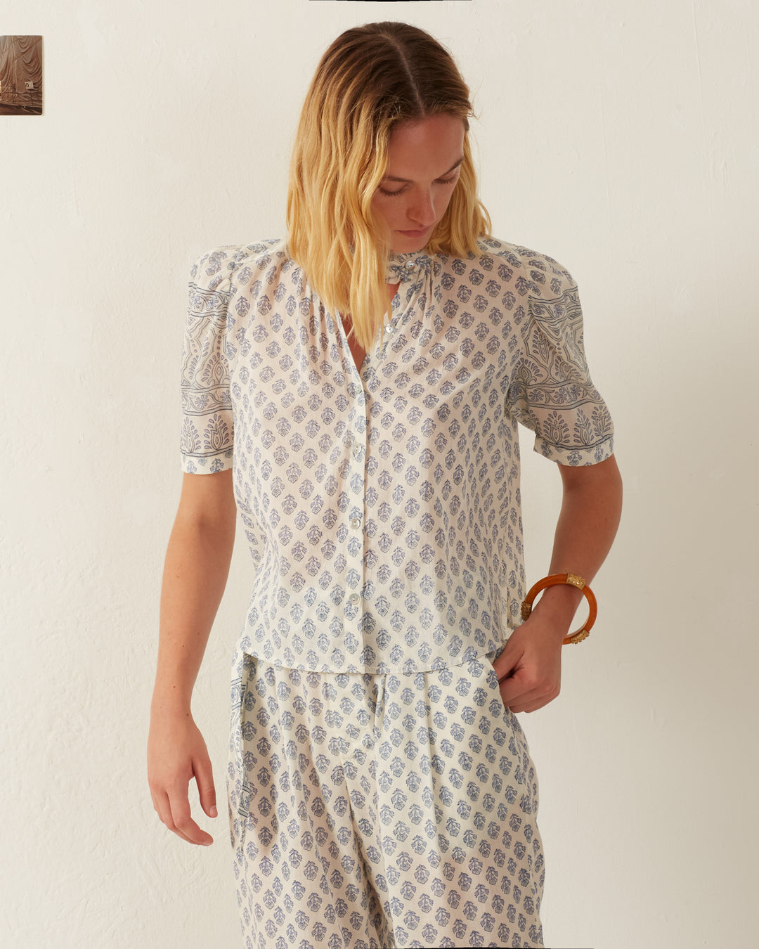 Wearing the Winnie Delft Bhutti Shirt, a vintage-inspired blue floral print made of cotton voile, the person gazes down to the left against an off-white background, accessorized with a brown bangle on their right wrist.