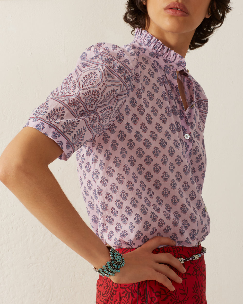 A person is sporting a Winnie Lilac Bhutti Shirt with intricate patterns and red pants. They have a turquoise bracelet on their wrist, all set against a plain white wall.