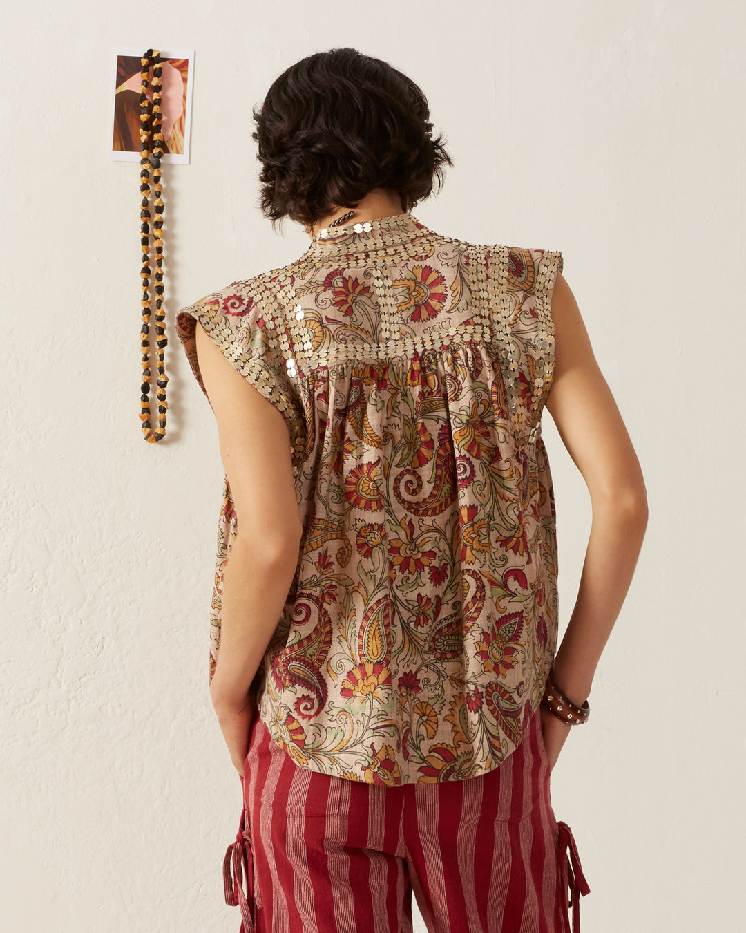 A person with short, dark hair stands facing a wall, wearing the Nehru Silver Terra Top adorned with floral and paisley designs. They are also in red and white striped pants. A photo and beaded necklace hang on the wall.