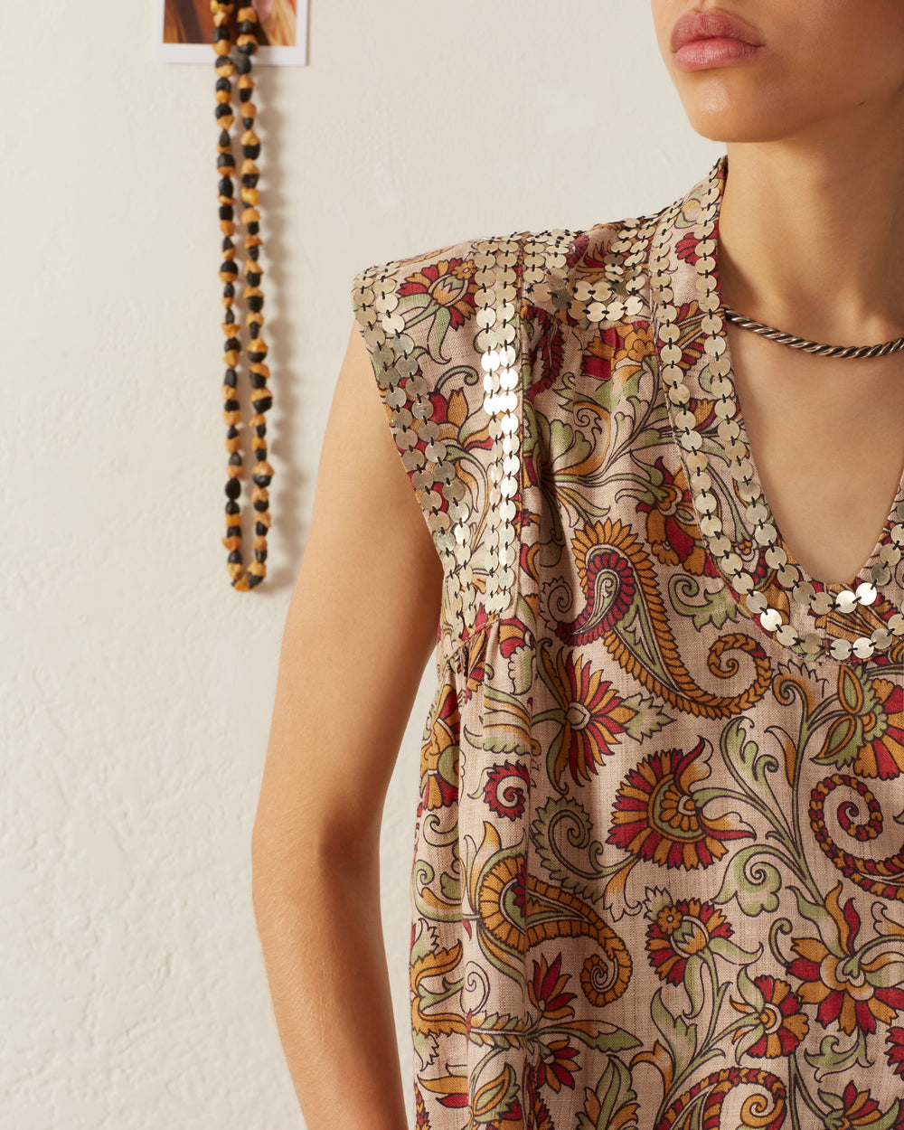 A person wearing the Nehru Silver Terra Top, featuring intricate floral patterns and silver paillettes, stands against a light-colored wall with a beaded necklace hanging in the background.