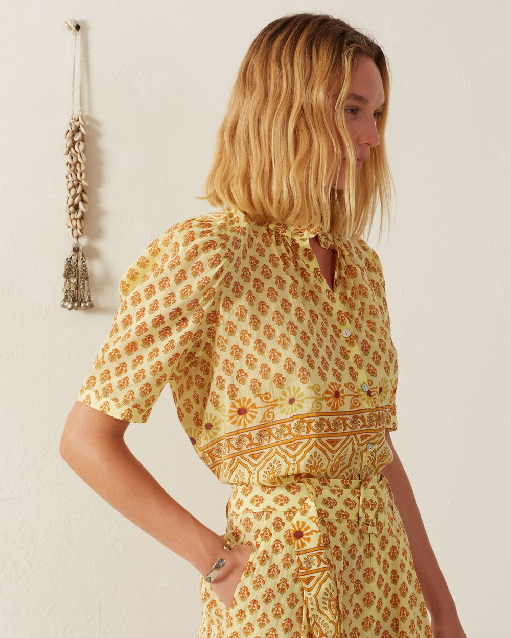 A person with shoulder-length blonde hair is wearing the Winnie Saffron Bhutti Shirt paired with matching pants, both adorned with intricate floral patterns. They stand in a softly lit room with a decorative tassel hanging on the cream-colored wall, creating a serene and vintage-inspired ambiance.