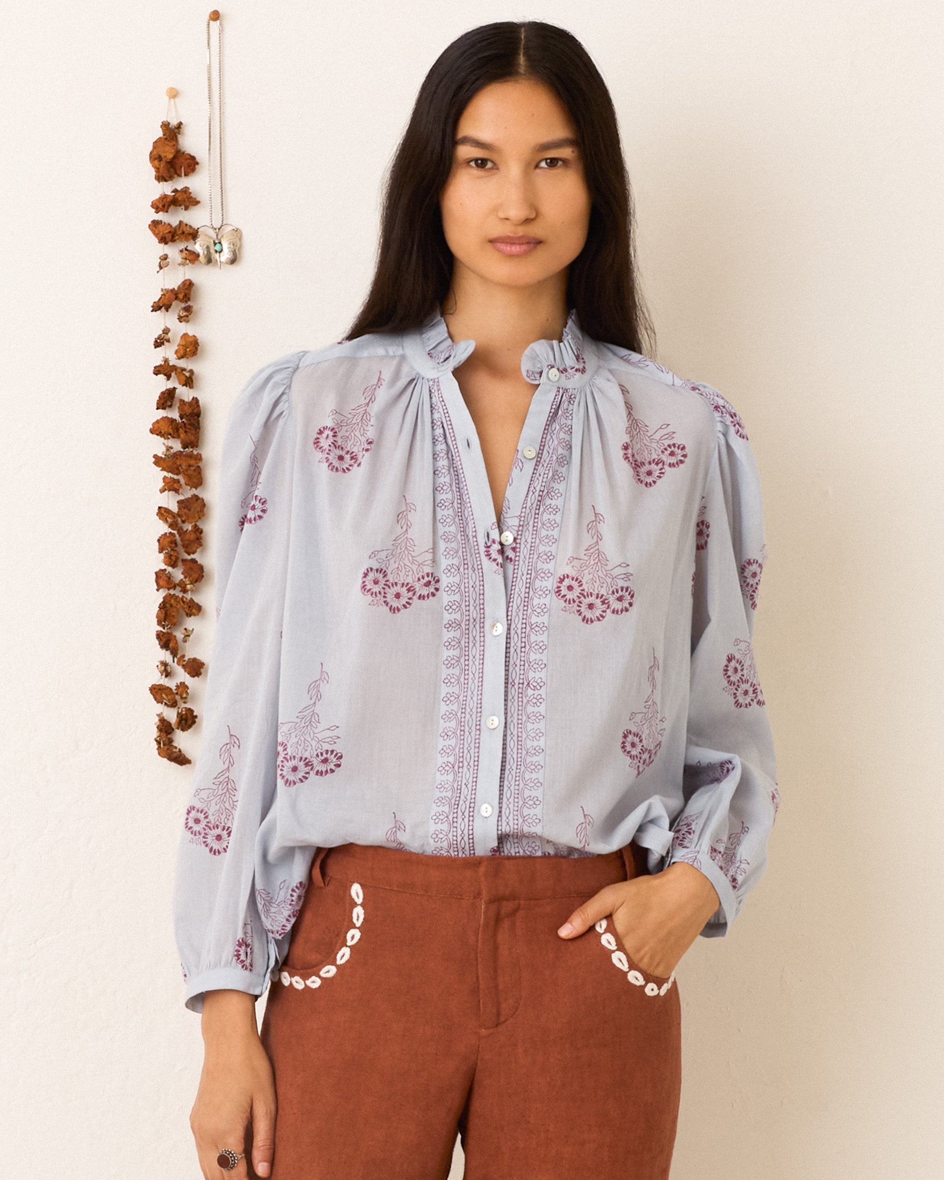 Wearing an Annabel Pansy Sky Shirt with brown pants, a person with long hair stands against a white wall decorated with a hanging dried plant.