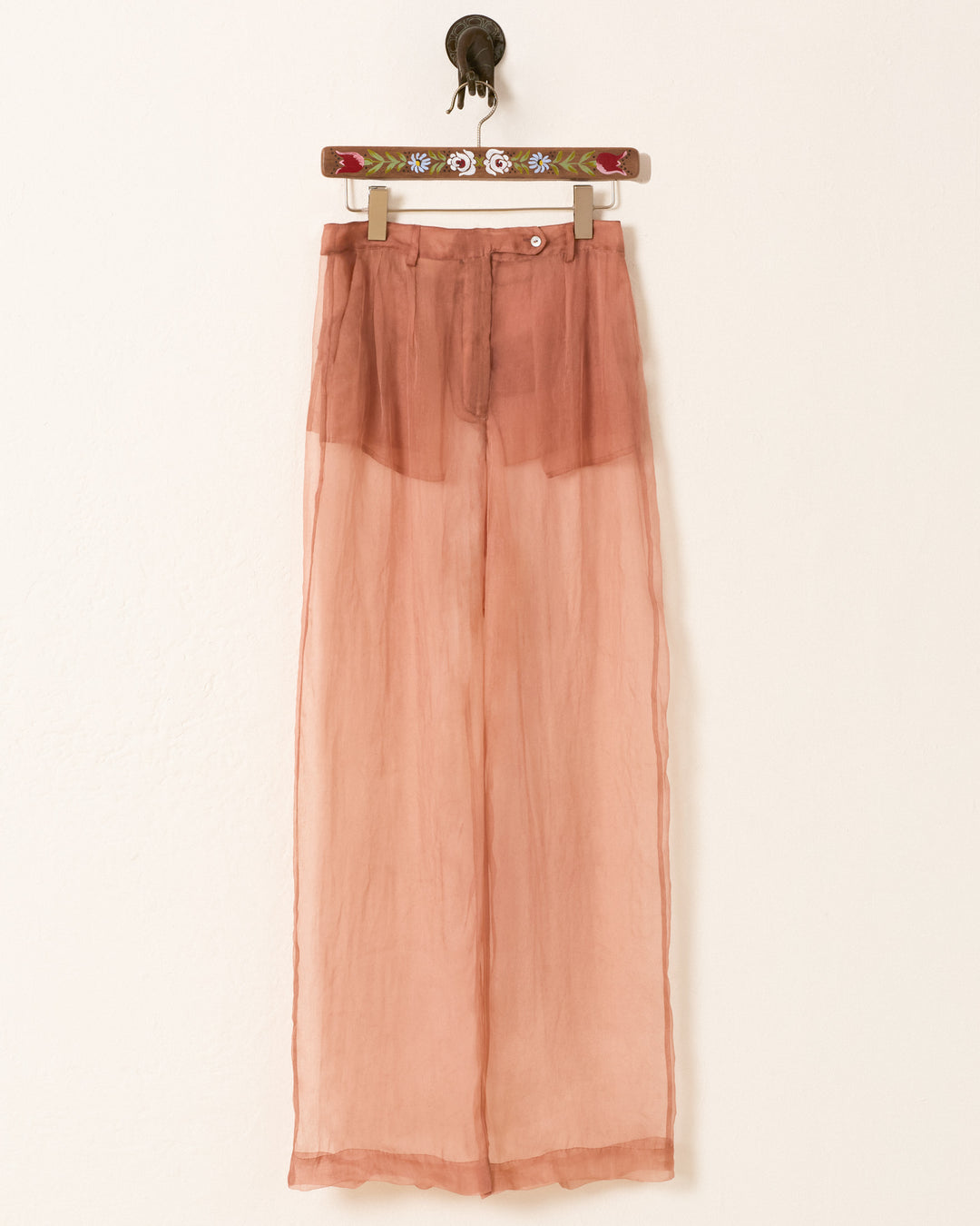 The Diana Mocha Organza Pant, featuring a sheer high-waisted design and wide-leg silhouette in peach, is displayed on a decorative hanger against a plain light wall.