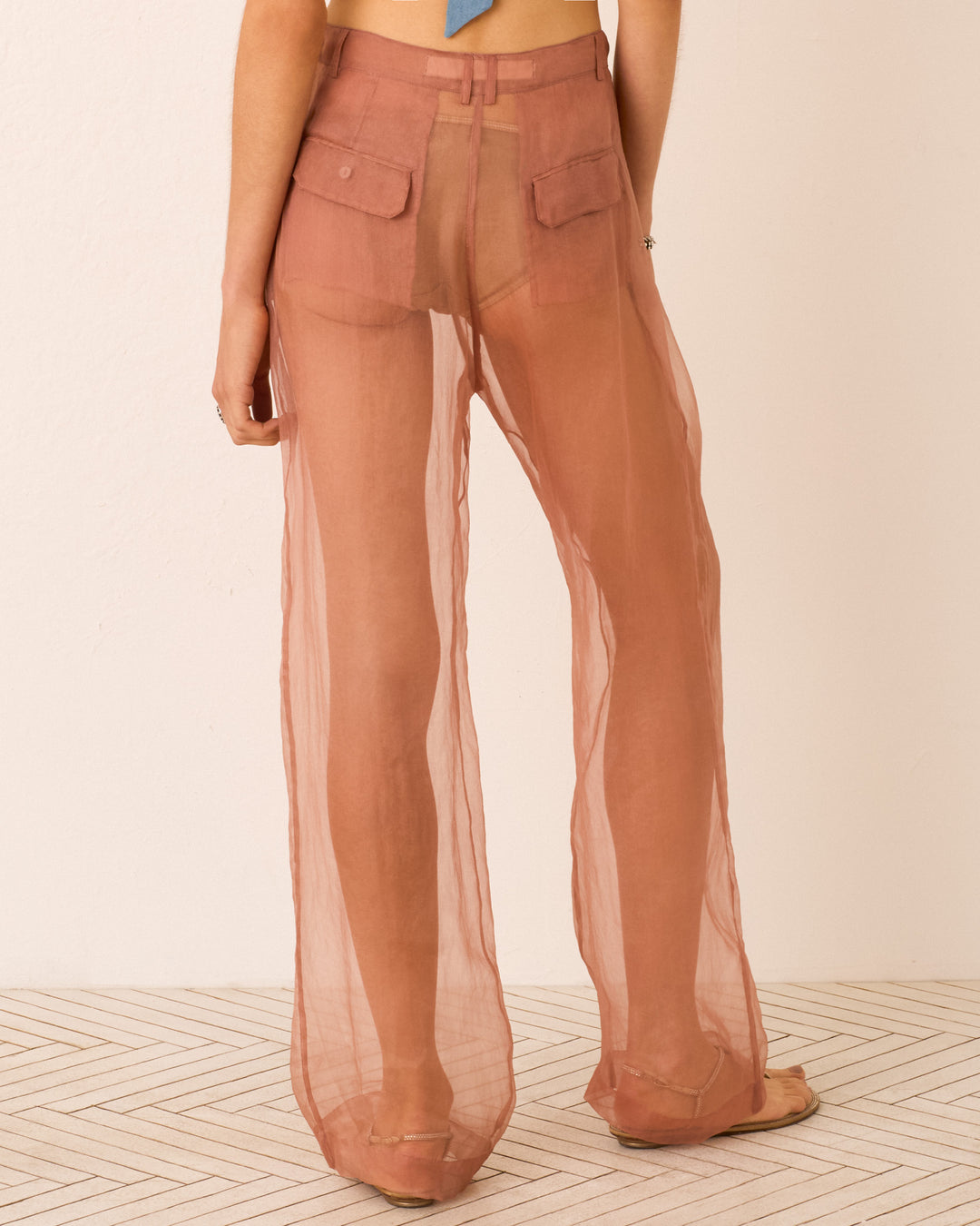 Person wearing Diana Mocha Organza Pant—sheer, high-waisted rust pants revealing underwear beneath—stands on a patterned floor, facing away from the camera.