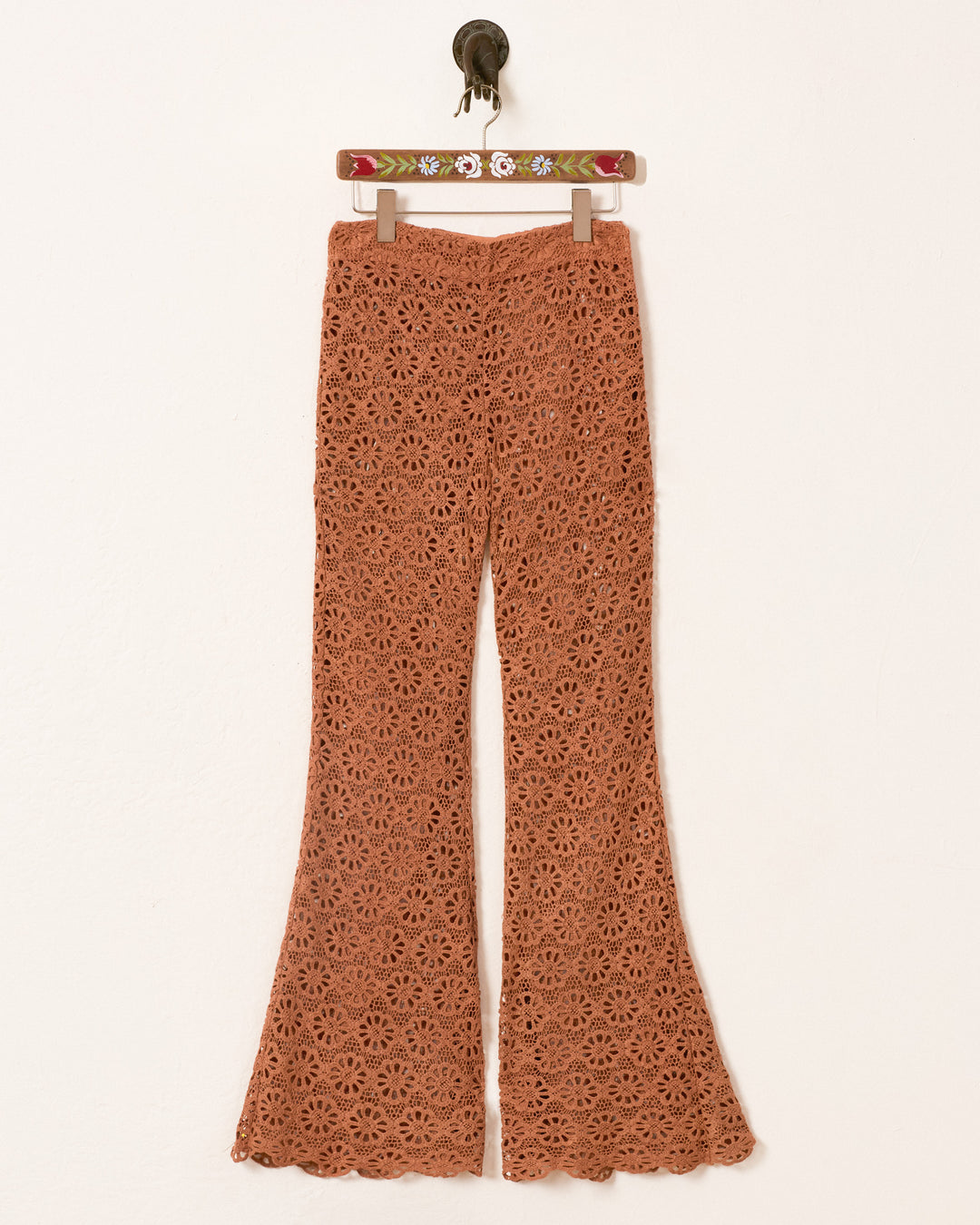 The Farah Mocha Daisy Pant—rust-colored, crochet bell-bottom flares with a daisy motif—hangs on a hanger against a plain wall.