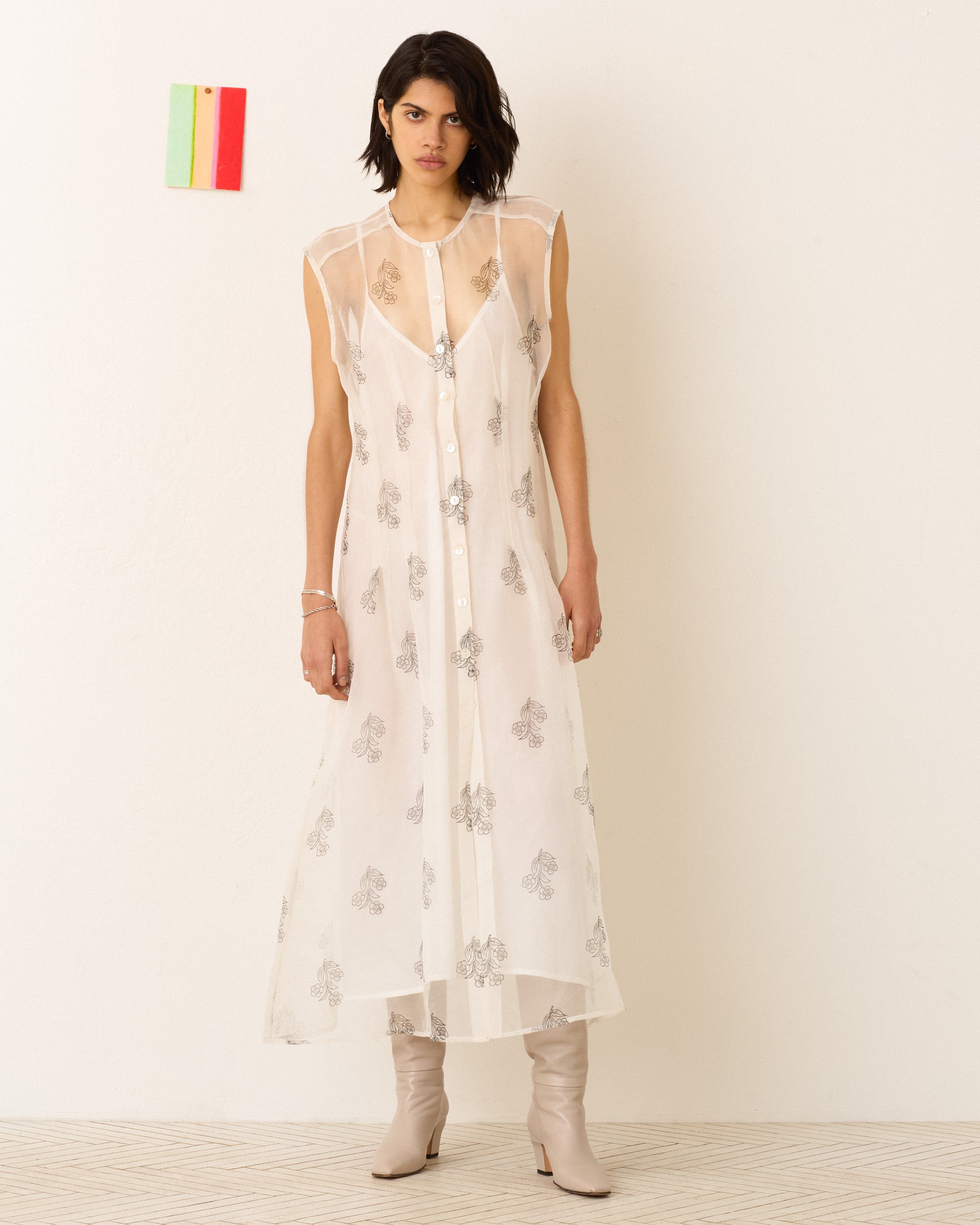 A woman with short dark hair wears the Gigi Lunar Flower Dress—a sheer white silk-organza piece with silver patterns—paired with beige boots, standing against a plain wall with a small, colorful object hanging behind her.