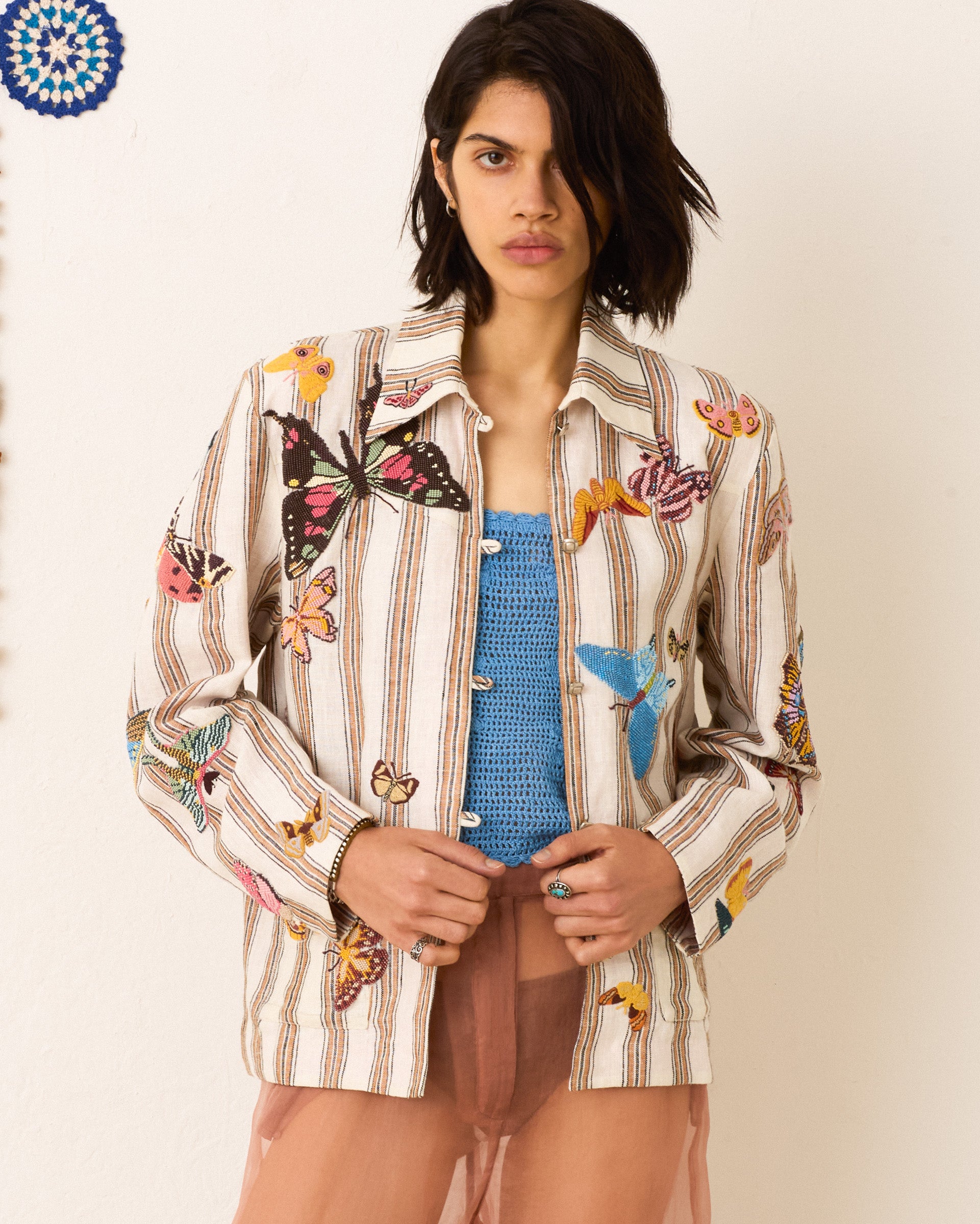 A person wearing the Jude Desert Butterfly Jacket, a vintage-inspired striped piece with colorful butterfly patterns, over a blue top and a translucent peach skirt stands against a light background.